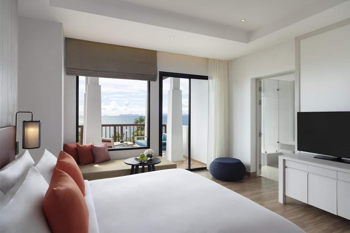 Habitación de hotel moderna con una cama grande, televisión, zona de estar con vista al mar y un balcón con puerta de vidrio.