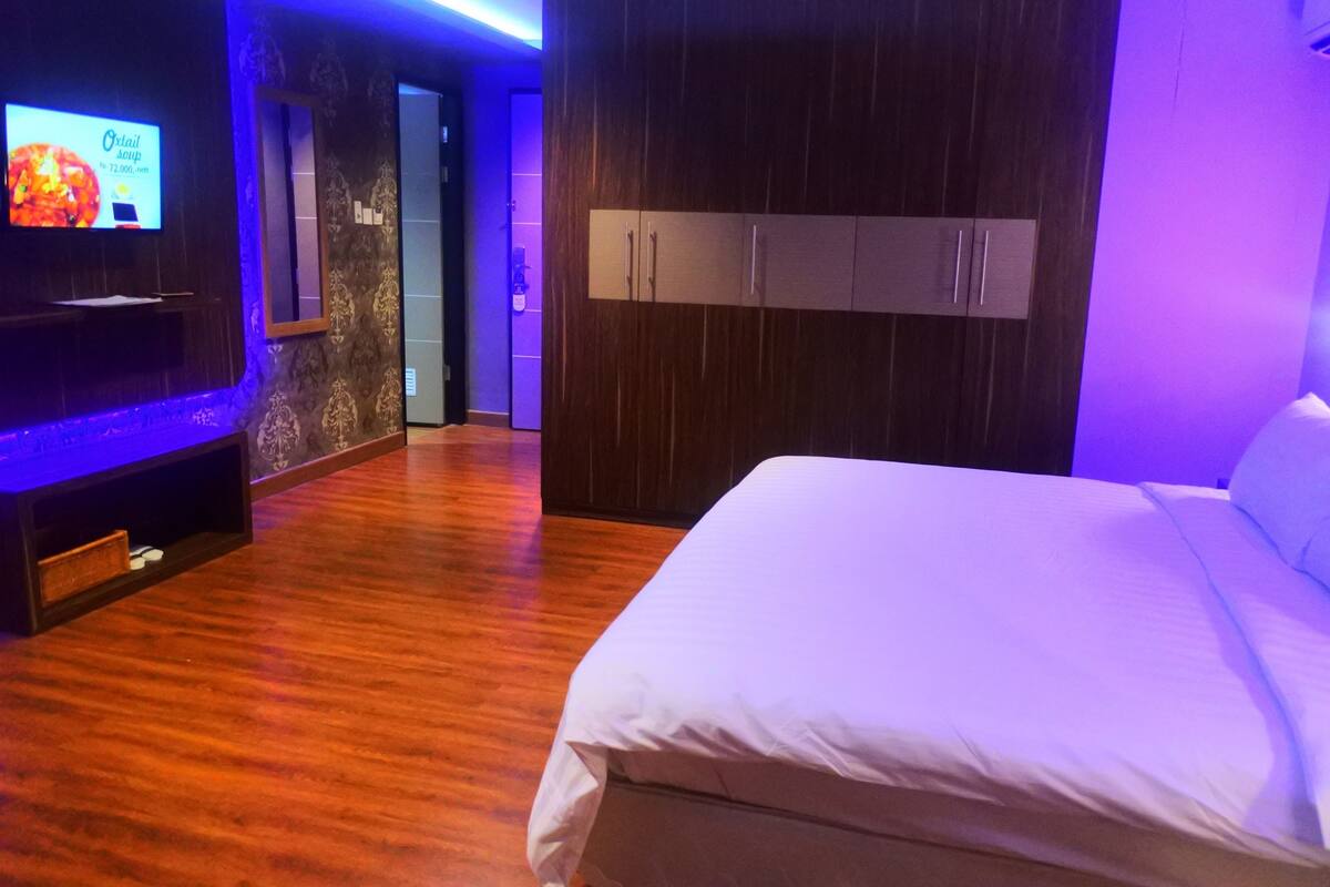 A hotel room with a bed, wooden flooring, a TV on the left wall, and a wardrobe with two doors.