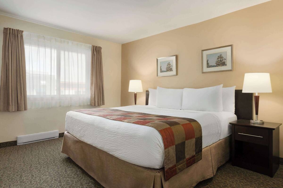 A hotel room with a large bed, two bedside tables with lamps, a window with curtains, and two framed pictures on the wall.
