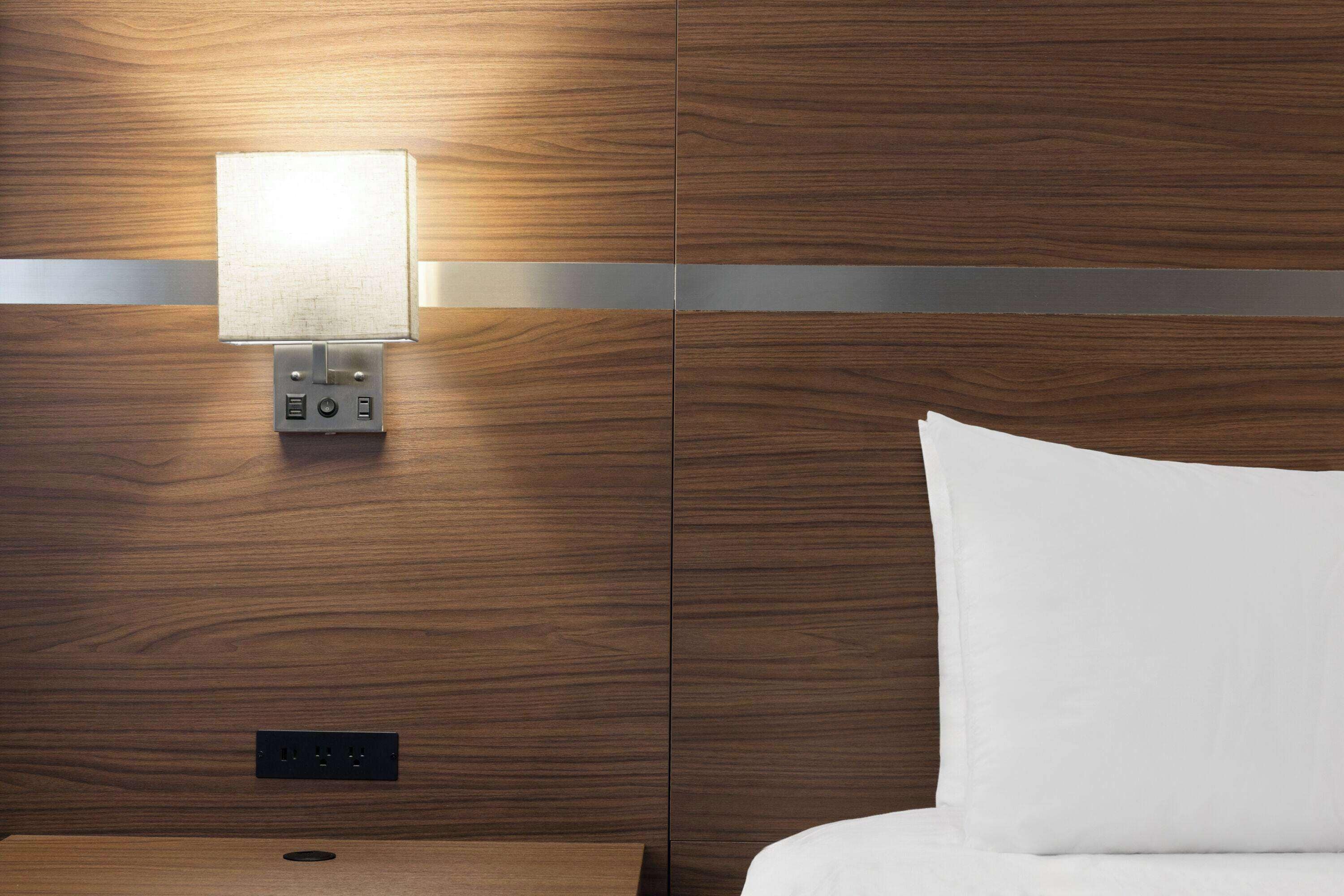 A hotel room with a wooden panel wall, a wall-mounted light, a power outlet, and a white bed.