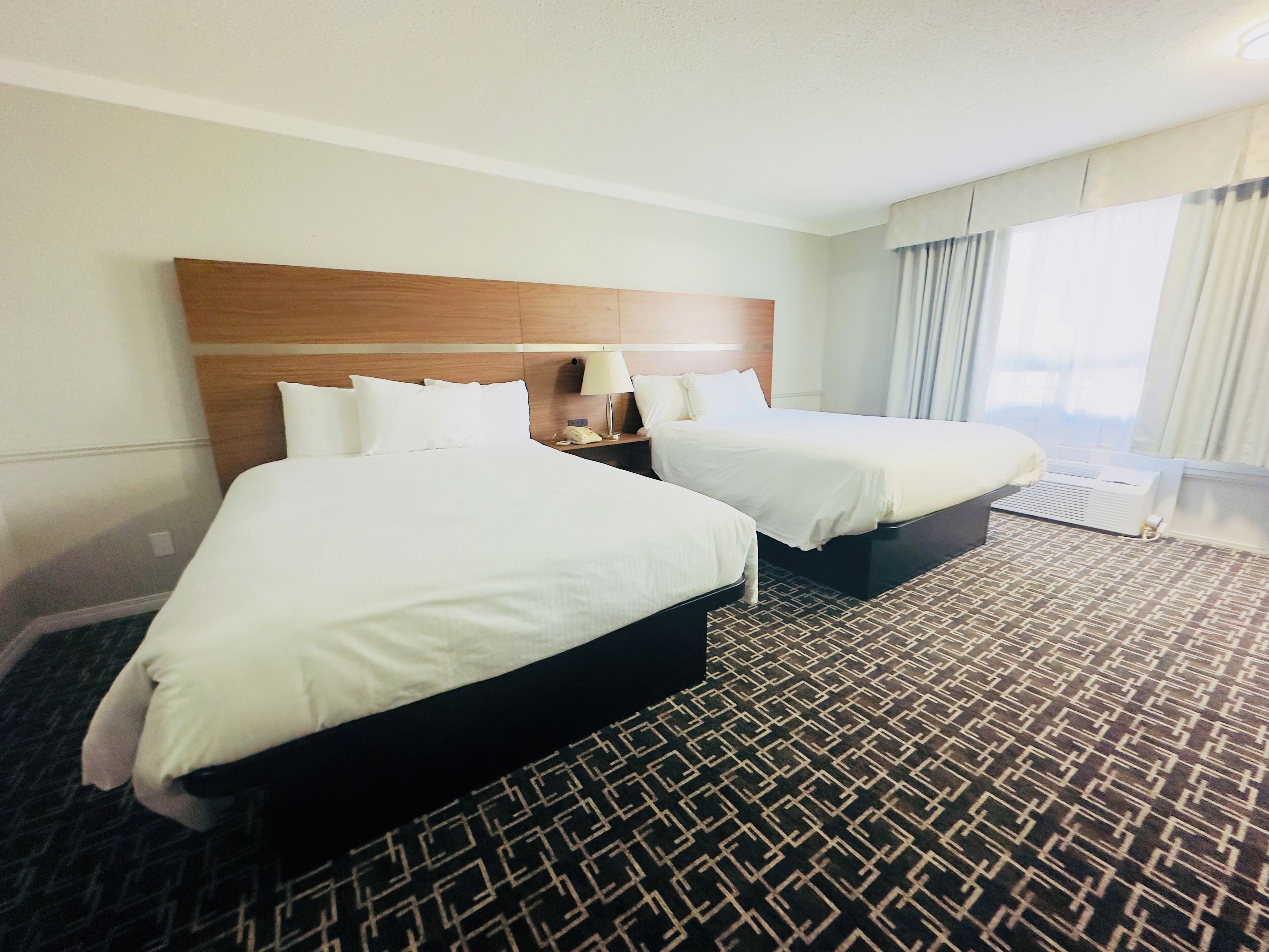 A hotel room with two beds, a wooden headboard, a nightstand with a lamp, and a patterned carpet.