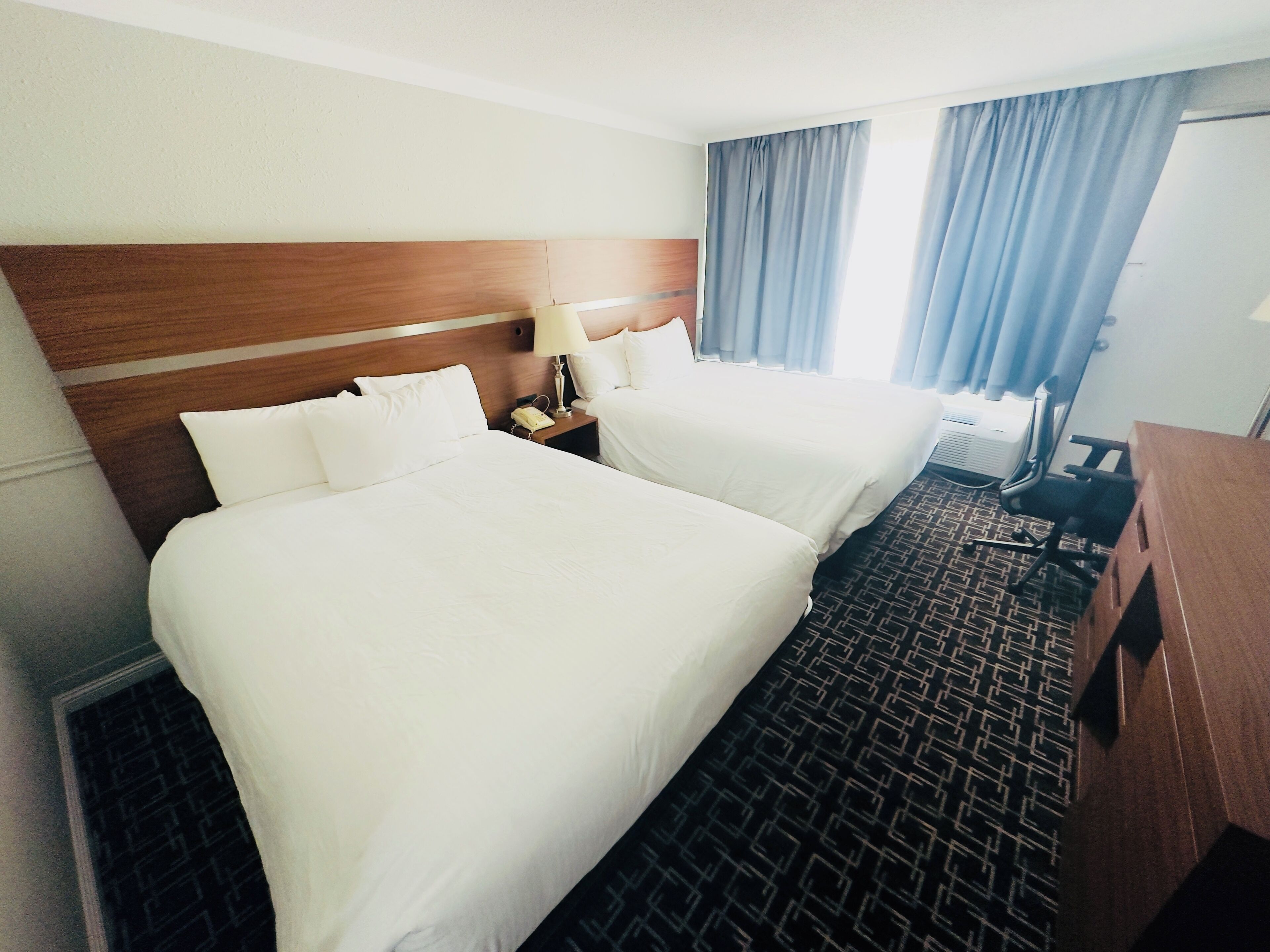 A hotel room with two beds, a wooden headboard, a nightstand with a lamp, a desk, and a patterned carpet.
