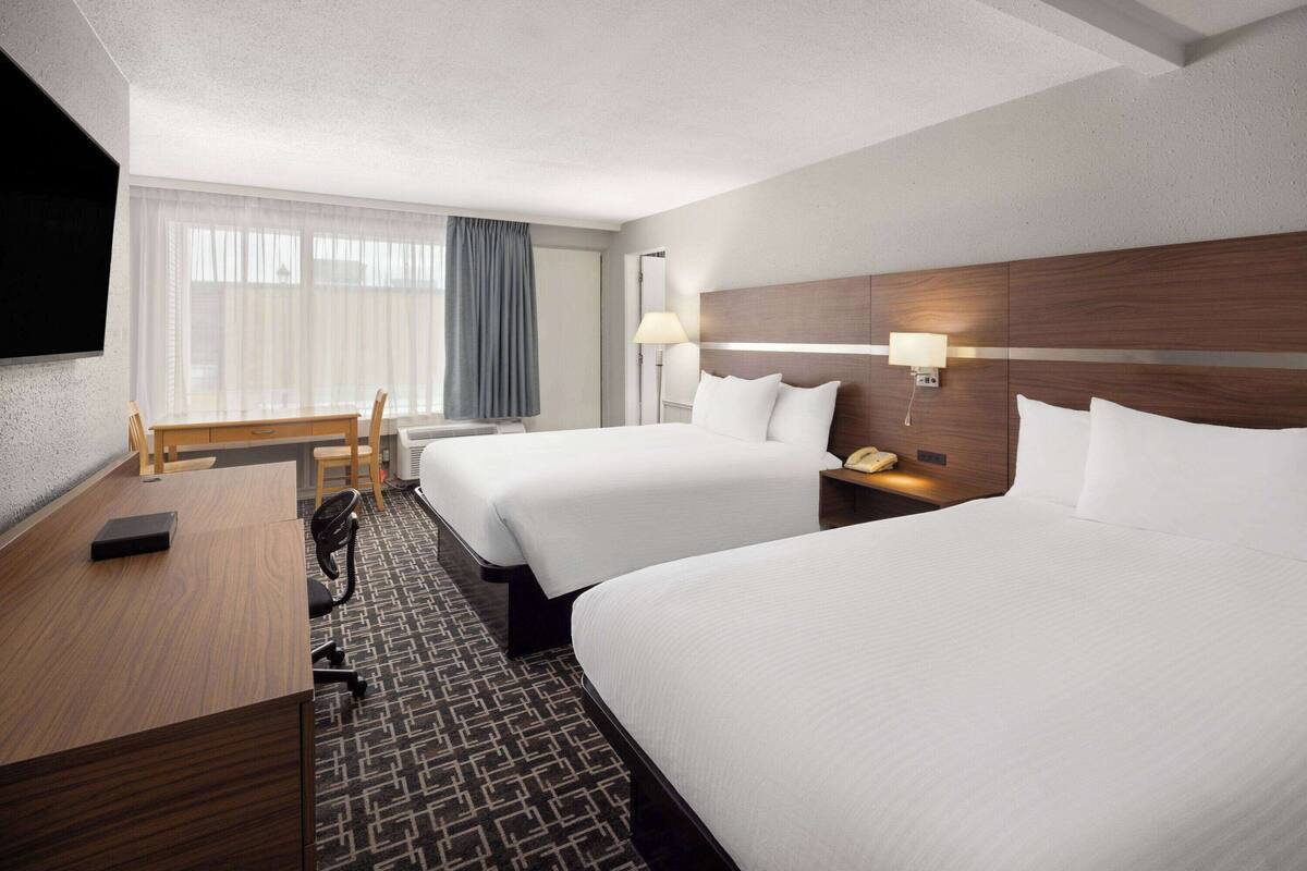 A hotel room with two beds, a desk, a chair, a TV, and a lamp.