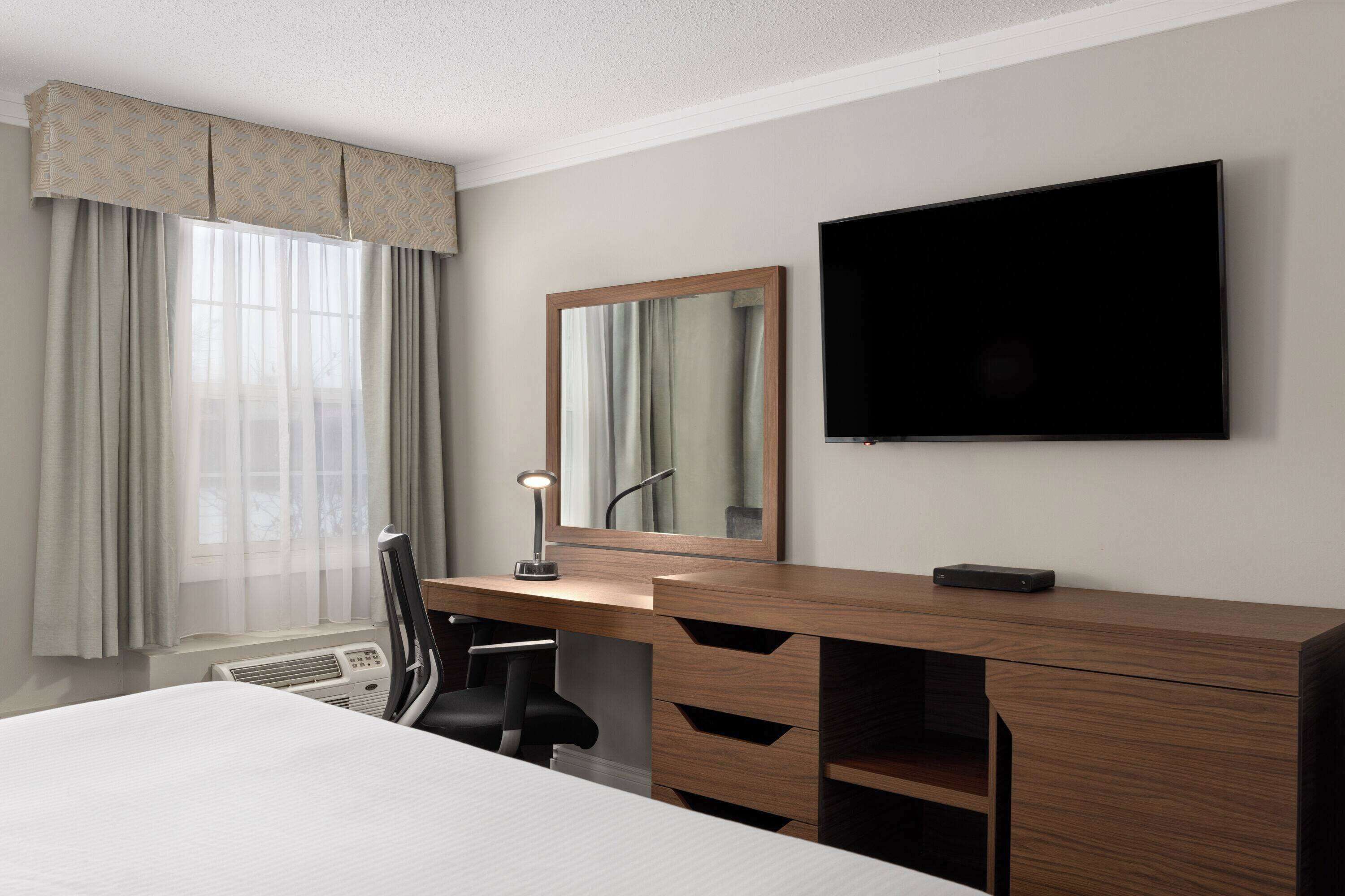 Una habitación de hotel con cama, escritorio con silla, lámpara de escritorio, televisor de pantalla plana y un ventanal con cortinas.
