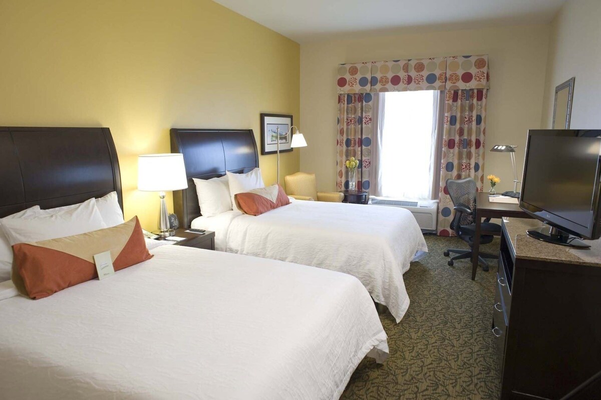 A hotel room with two beds, a desk, a television, and a window with curtains.