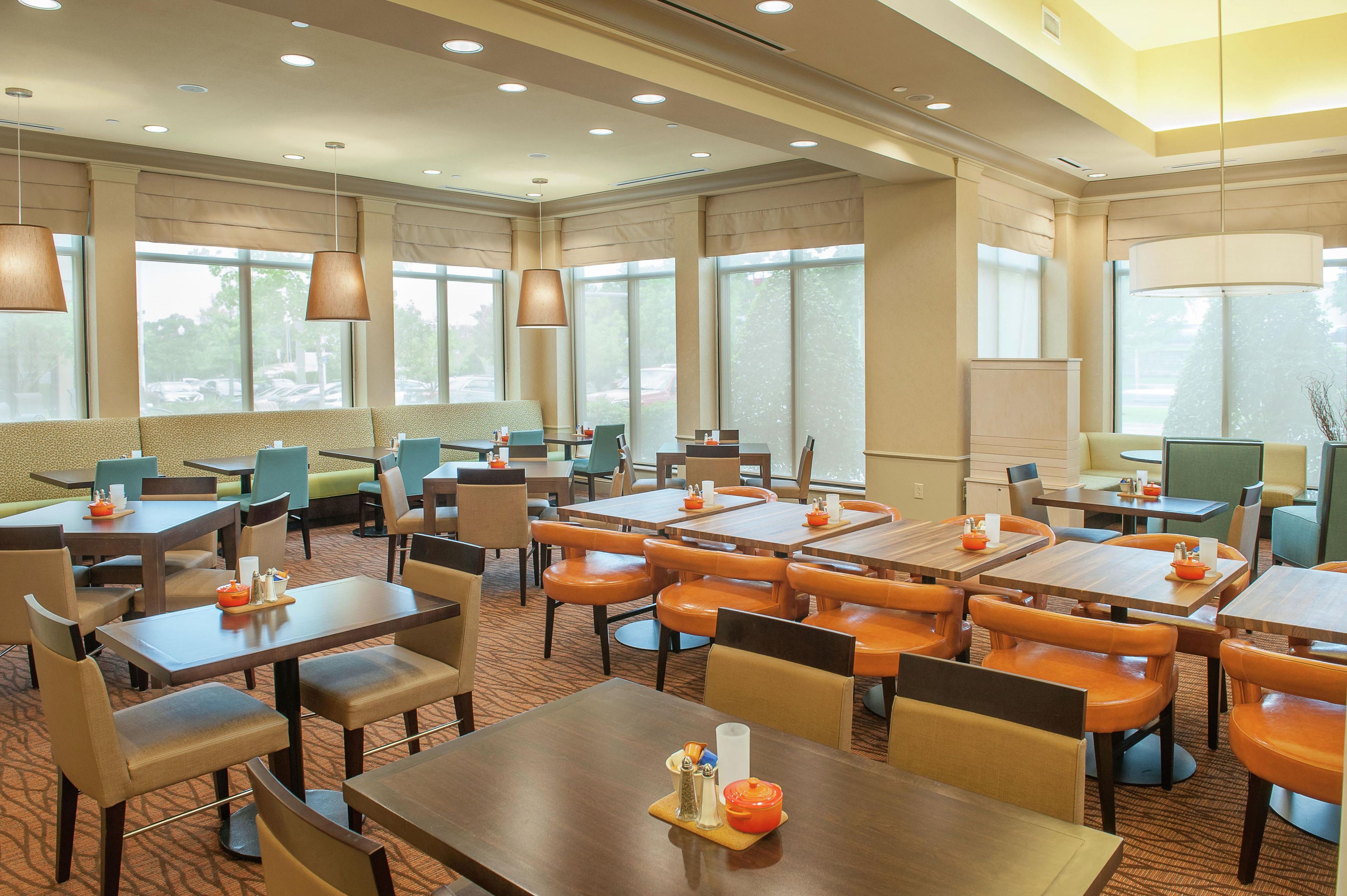 A hotel dining area with multiple tables and chairs, large windows, and ceiling lights.