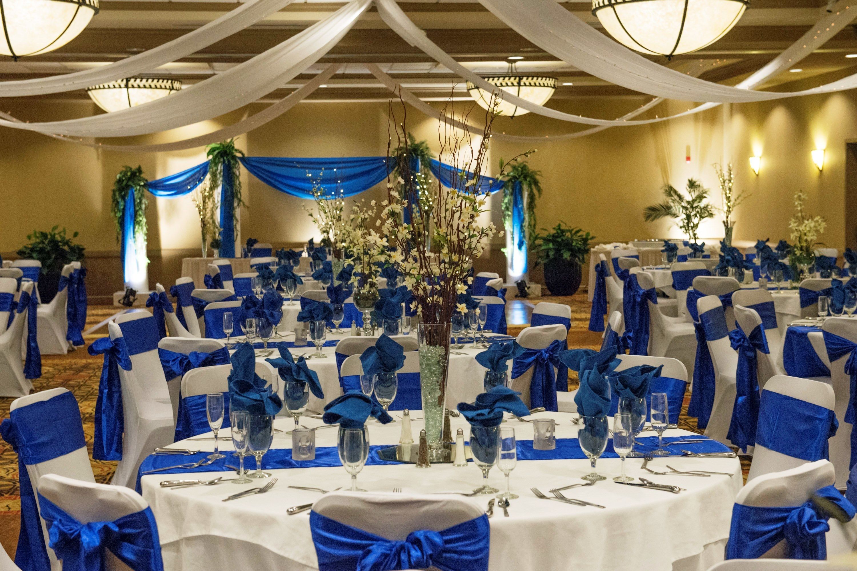 Uma sala de banquetes com mesas redondas preparadas para um evento formal, com decoração azul e branca.