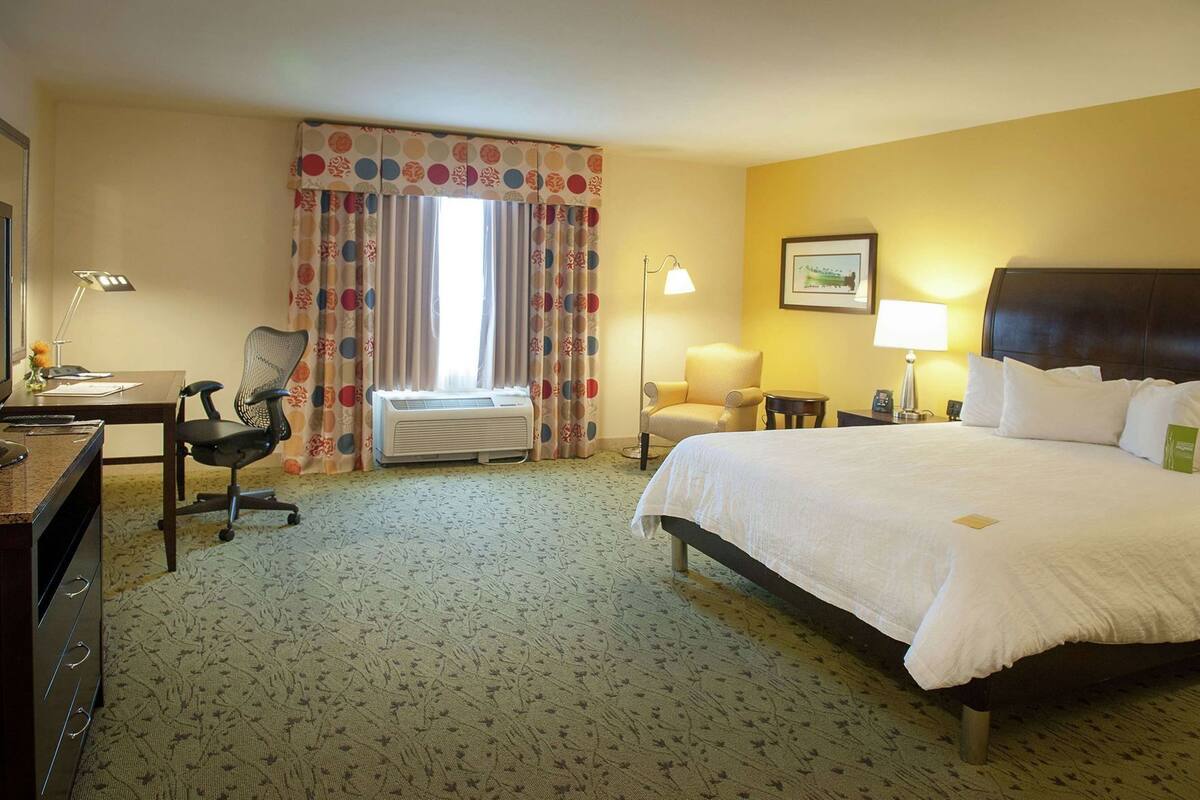 A hotel room with a bed, desk, chair, lamp, and air conditioning unit.