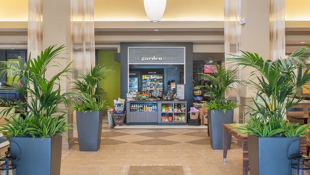 A spacious lobby with hanging pendant lights, large potted plants, and a visible store named "The Garden Market" in the background. - Hilton Garden Inn Pensacola Airport - Medical Center (Pensacola)