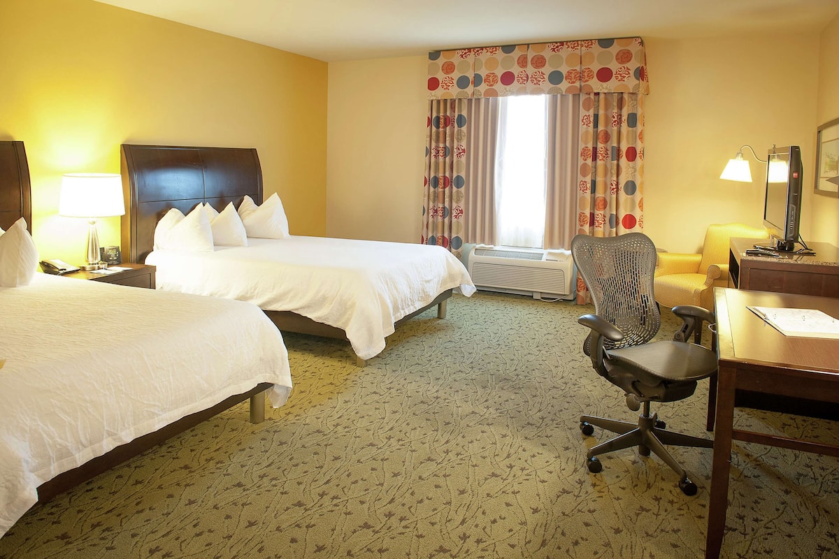 A hotel room with two beds, a desk with a chair, a lamp, and a window with curtains.