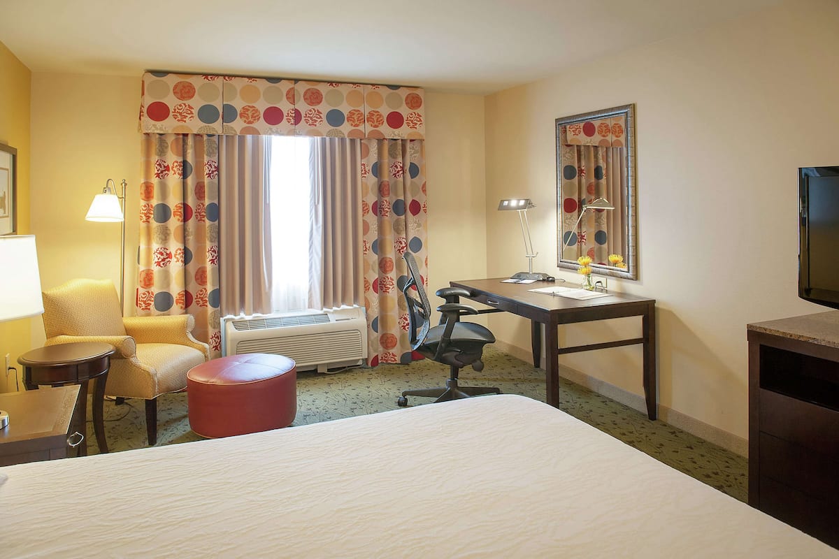 A hotel room with a bed, a desk, a chair, a red ottoman, a lamp, a mirror, and a television.