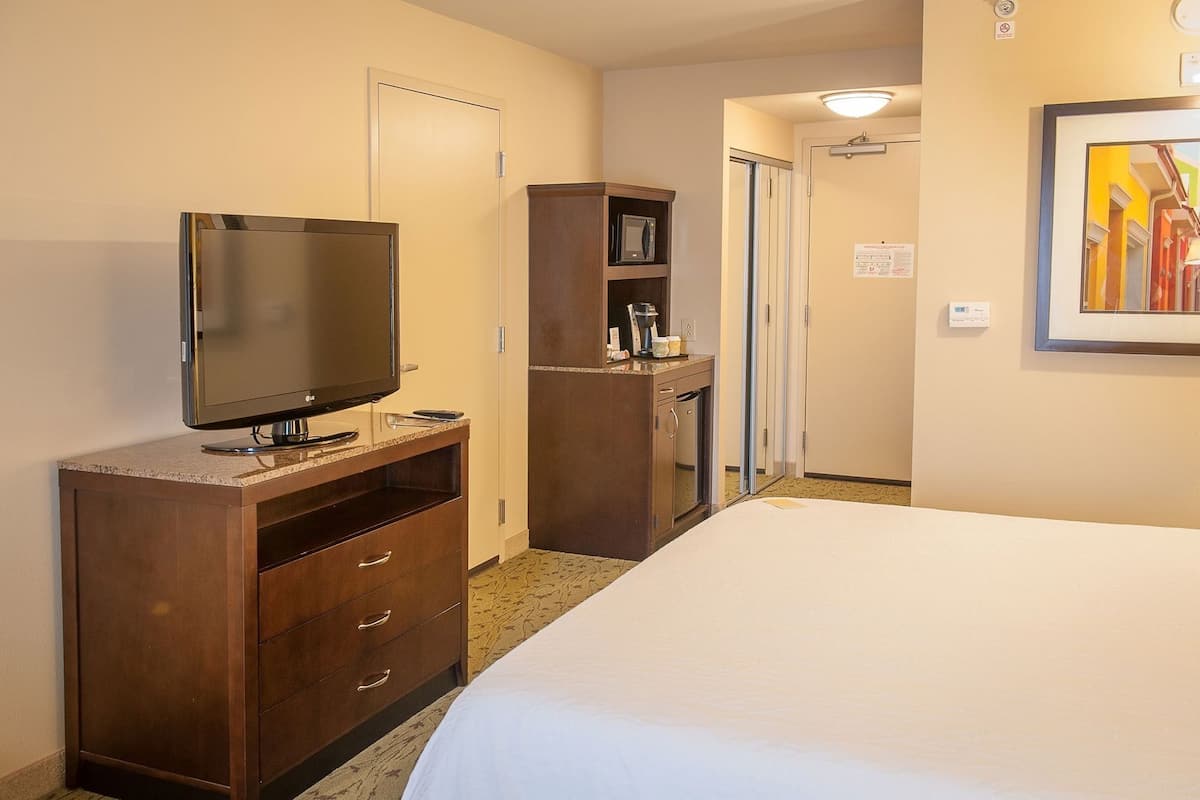 A hotel room with a bed, a TV on a wooden stand, a minibar, and a door leading to another room.
