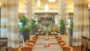 A spacious dining area with a long table, multiple chairs, and hanging pendant lights. There are potted plants and a central display case with items on the table. - Hilton Garden Inn Pensacola Airport - Medical Center (Pensacola)