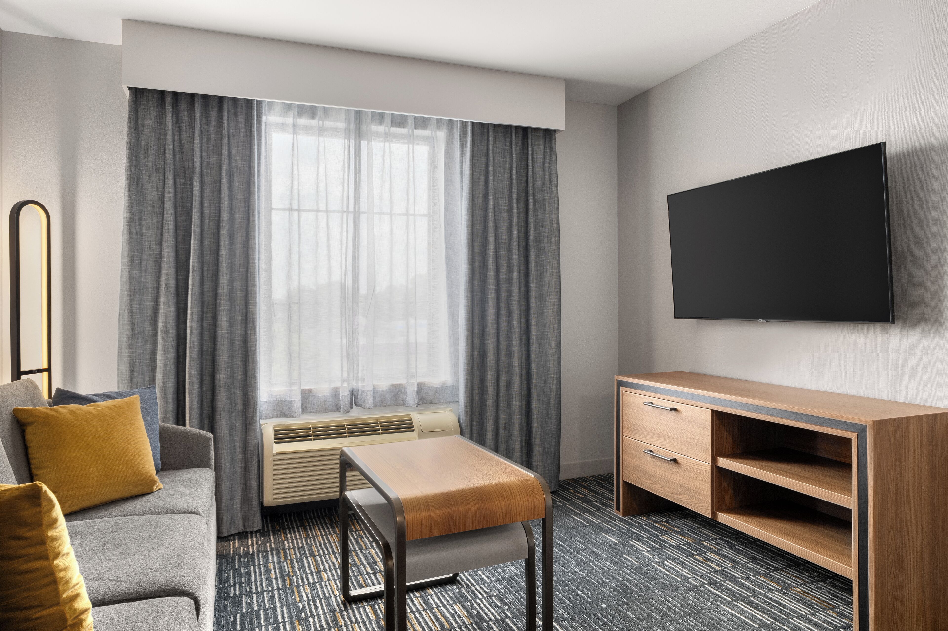 A hotel room with a sofa, a wooden table, a TV mounted on the wall, and a large window with curtains.