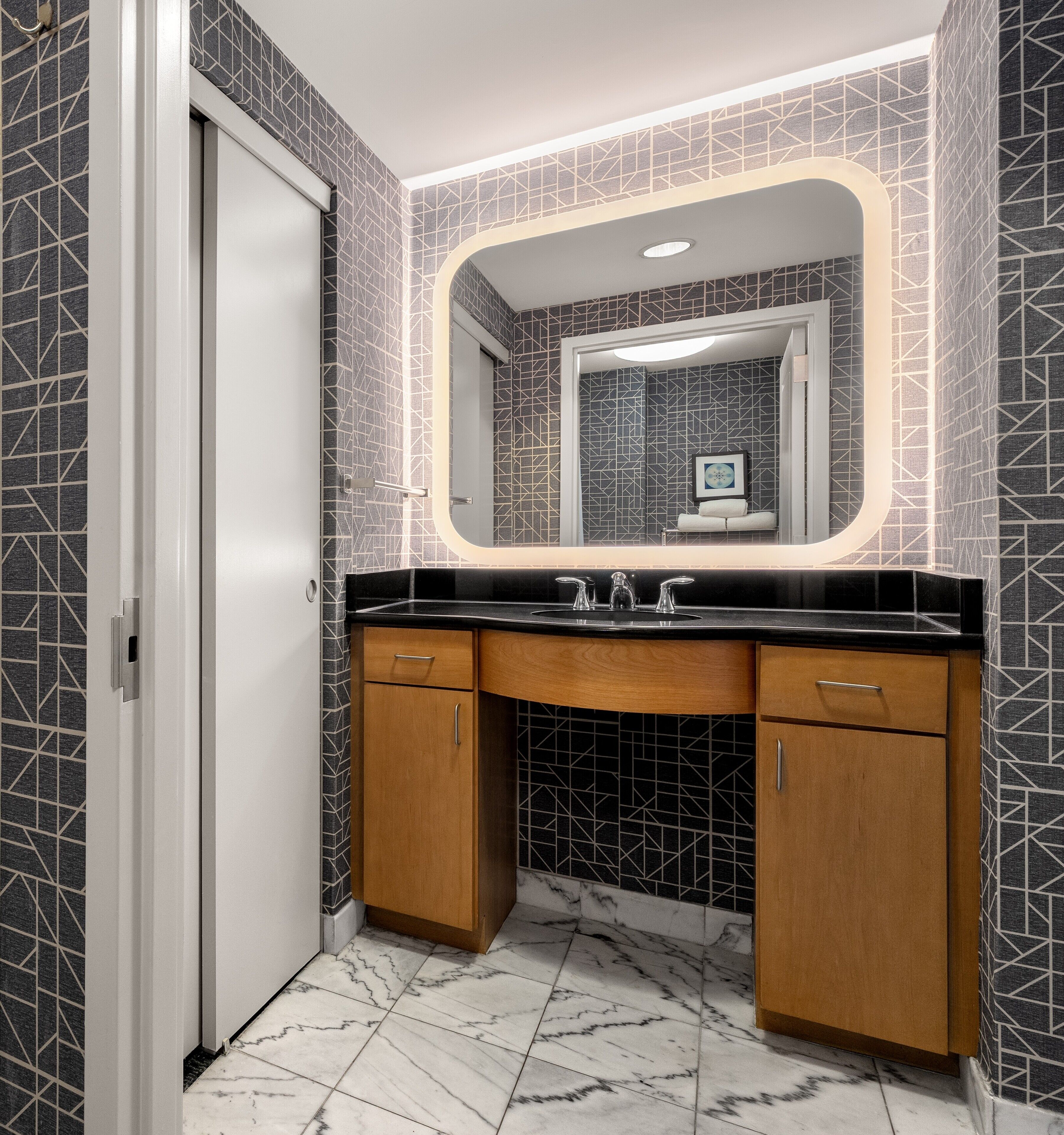 Une salle de bain avec un comptoir noir, des meubles en bois et un miroir agrémenté d’un pare-feu en carreaux aux motifs géométriques.