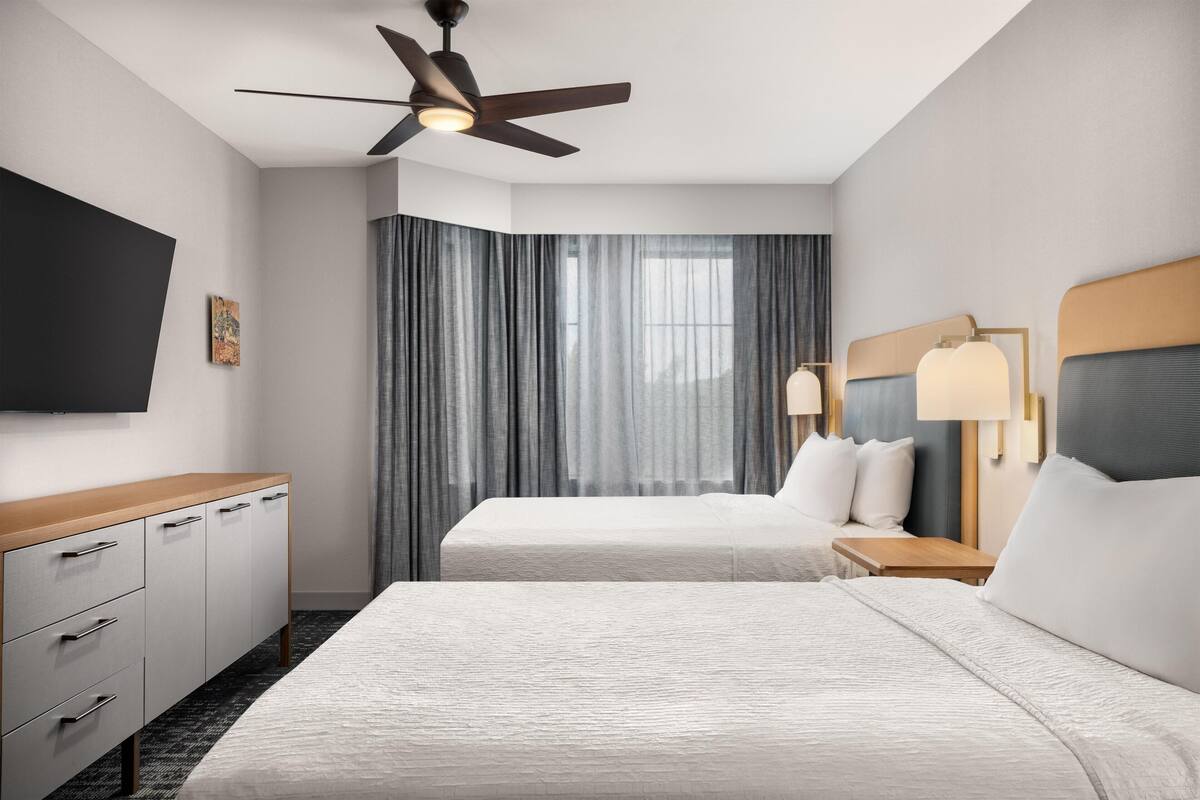 A hotel room with two beds, a ceiling fan, a flat-screen TV, a desk, and a window with curtains.