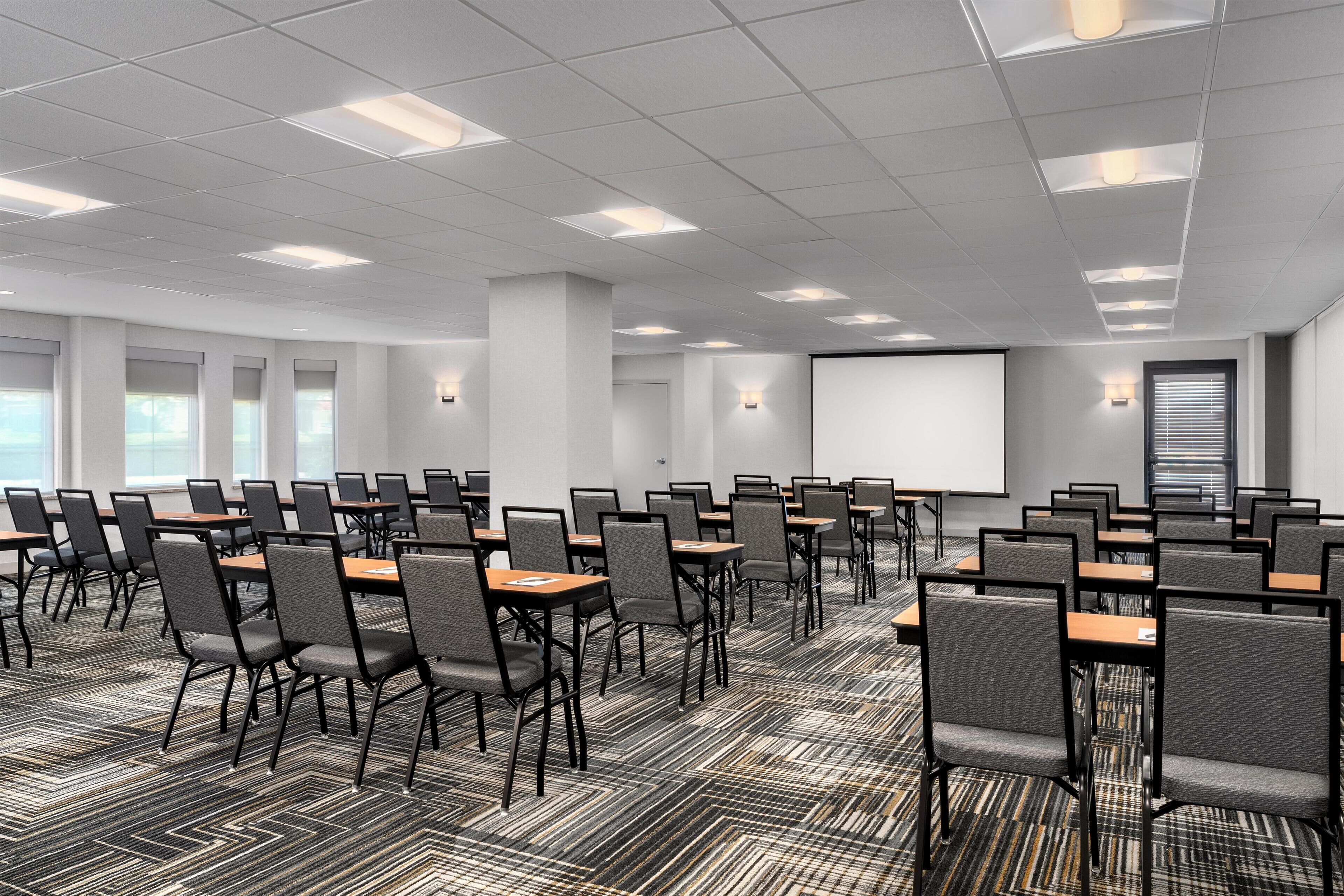 Une salle de conférence avec des rangées de tables et de chaises, un écran de projection et une grande fenêtre.