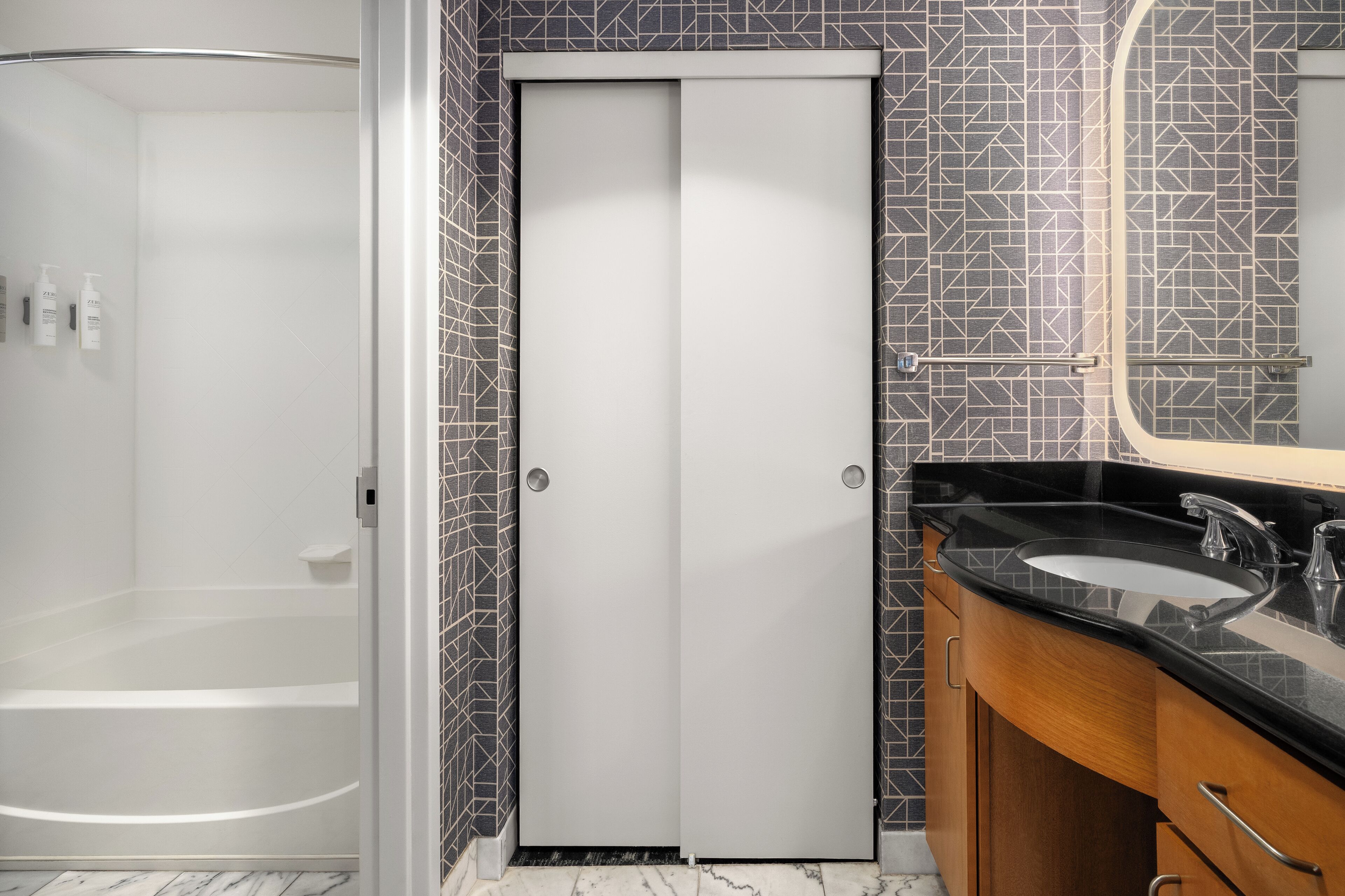 Une salle de bain avec une douche, une porte blanche, un lavabo avec un plan de travail noir et un miroir.