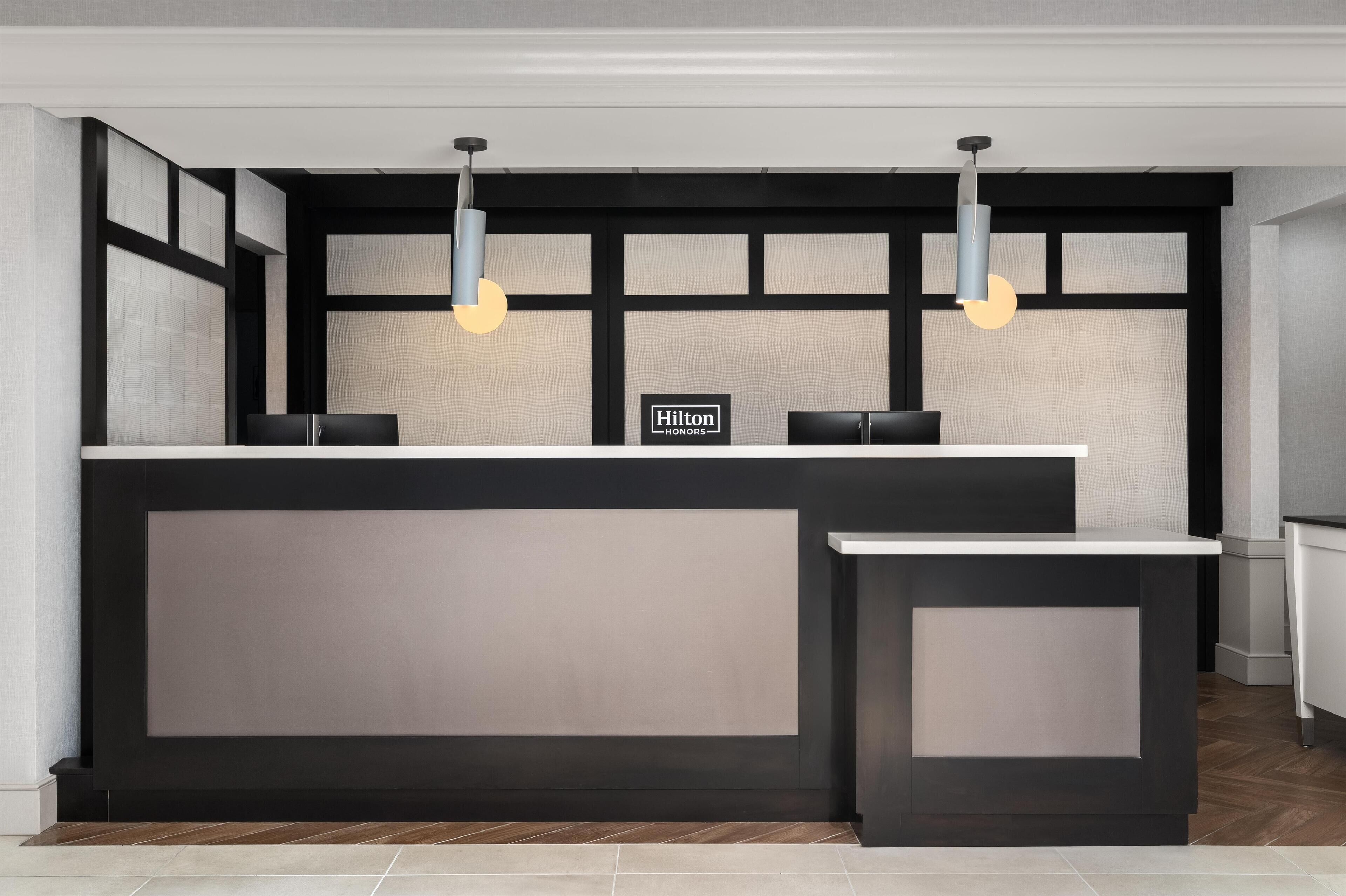 A modern hotel reception desk with a minimalist design, featuring two pendant lights and a sign indicating it is a Hilton Honors location.