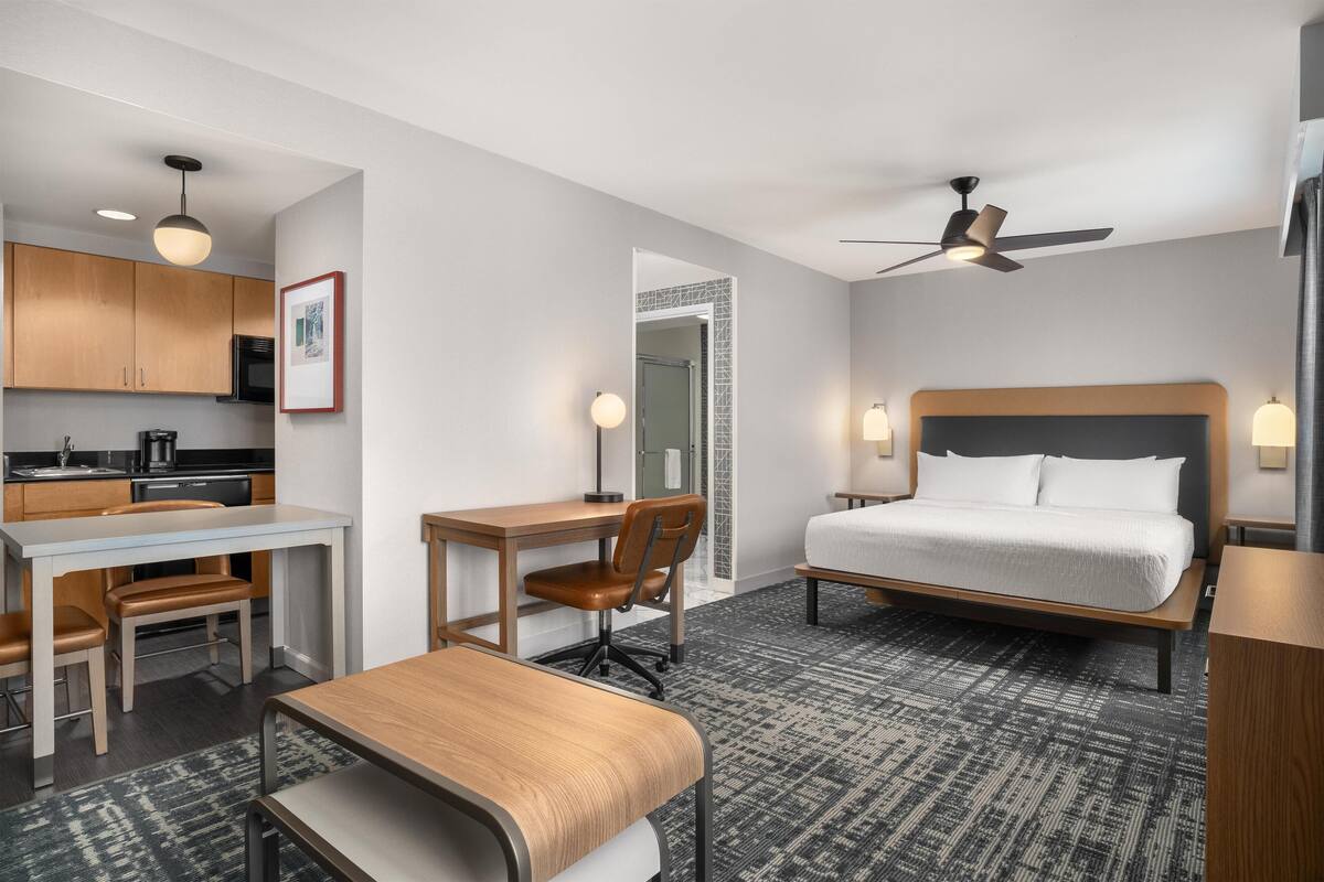 Habitación de hotel con cama, escritorio, silla, kitchenette y ventilador de techo.