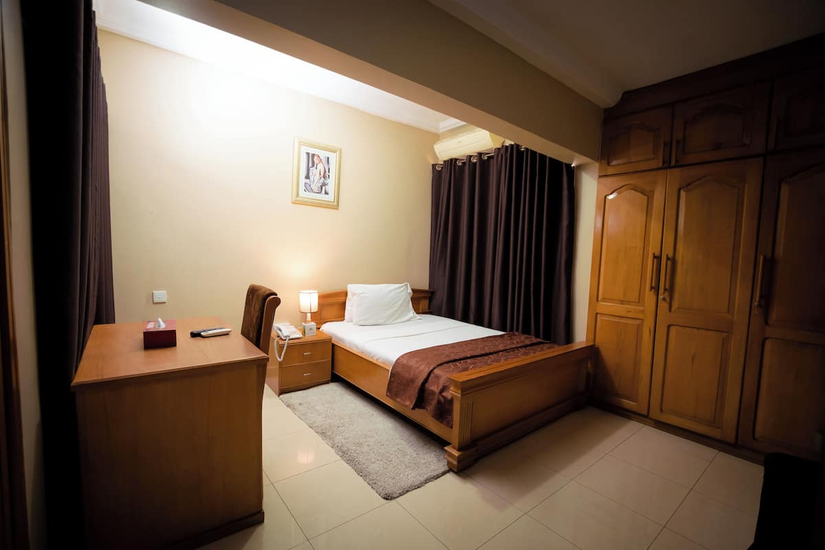 A neatly arranged hotel room with a bed, a desk with a lamp, a chair, and a wardrobe.