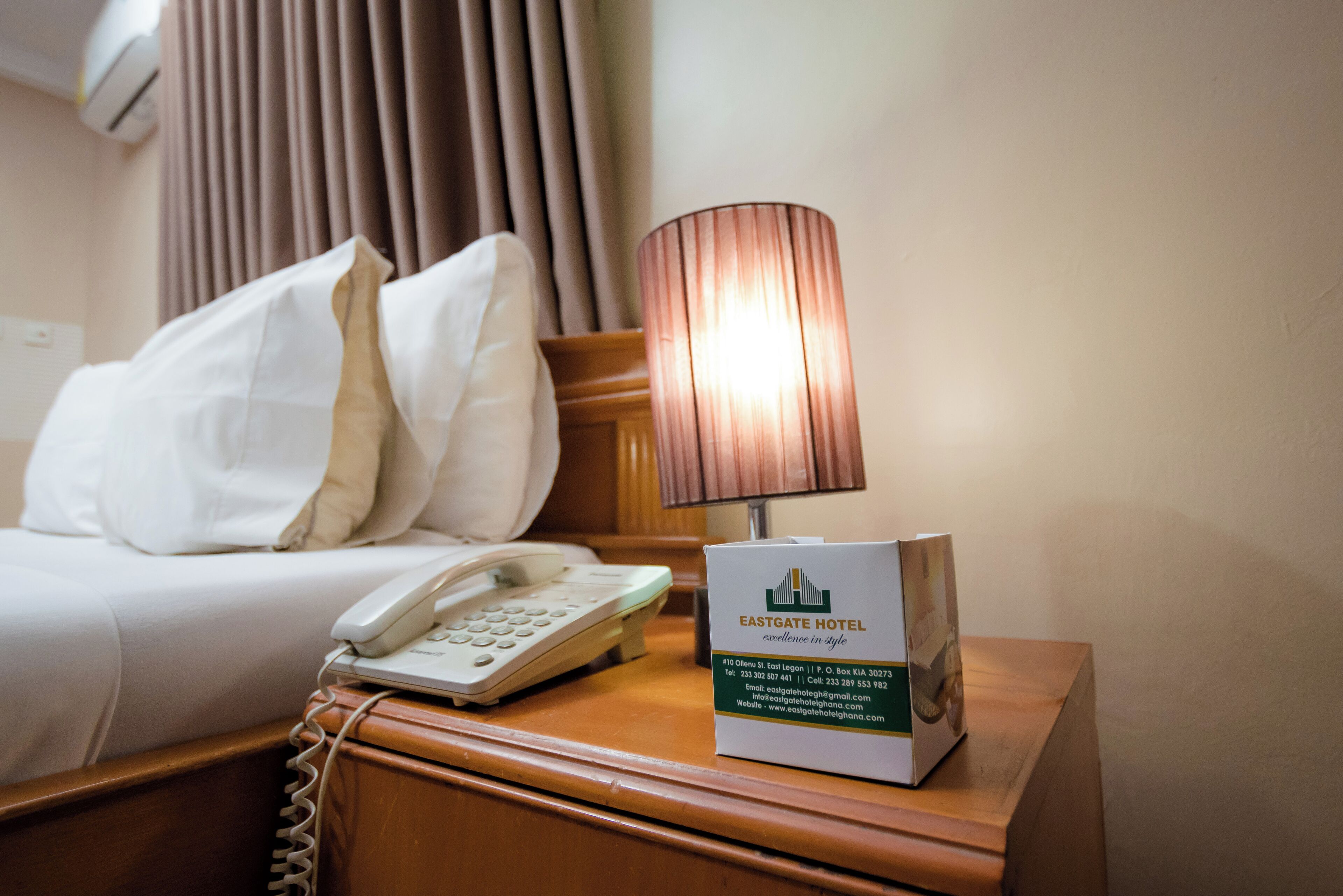 Un lit d’hôtel bien fait, avec une lampe et un téléphone sur une table de chevet en bois. On peut également voir une enseigne indiquant le nom de l’hôtel et ses coordonnées.