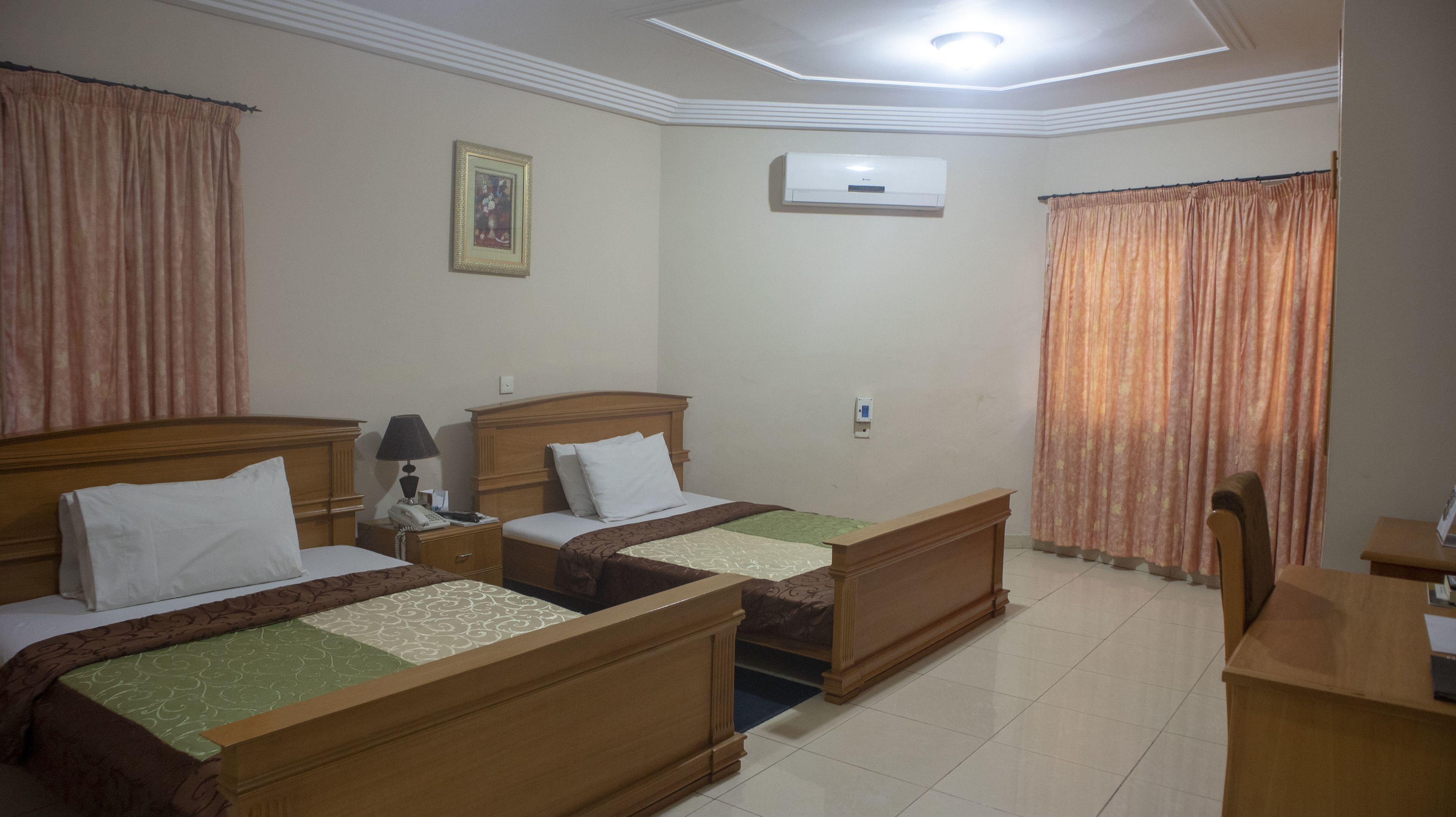 Une chambre d’hôtel avec deux lits en bois, un bureau, une chaise, un appareil de climatisation et un téléphone sur la table de chevet.
