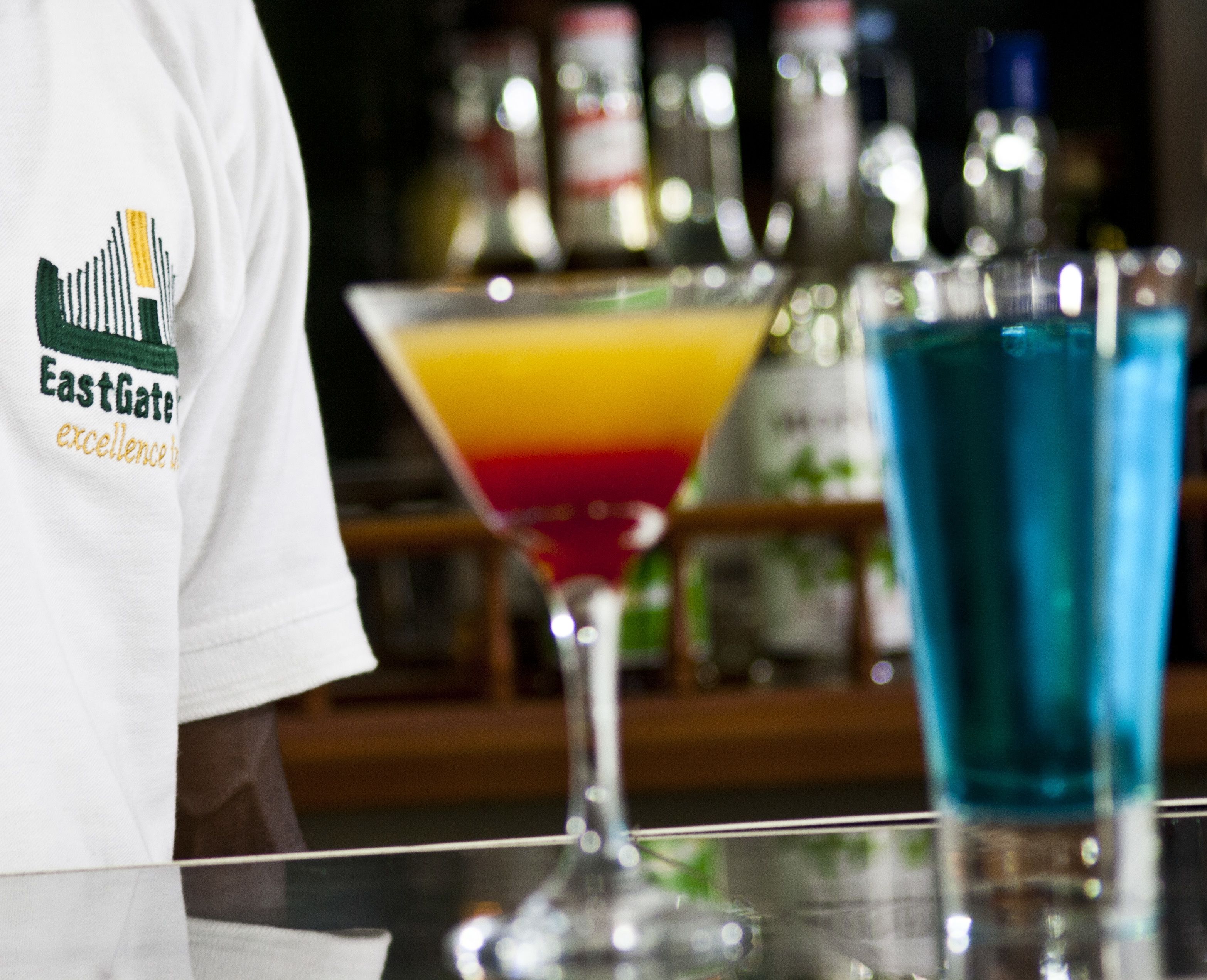 A bar area with a bartender wearing a white shirt with a logo, a colorful drink in a martini glass, and a blue drink in a glass.
