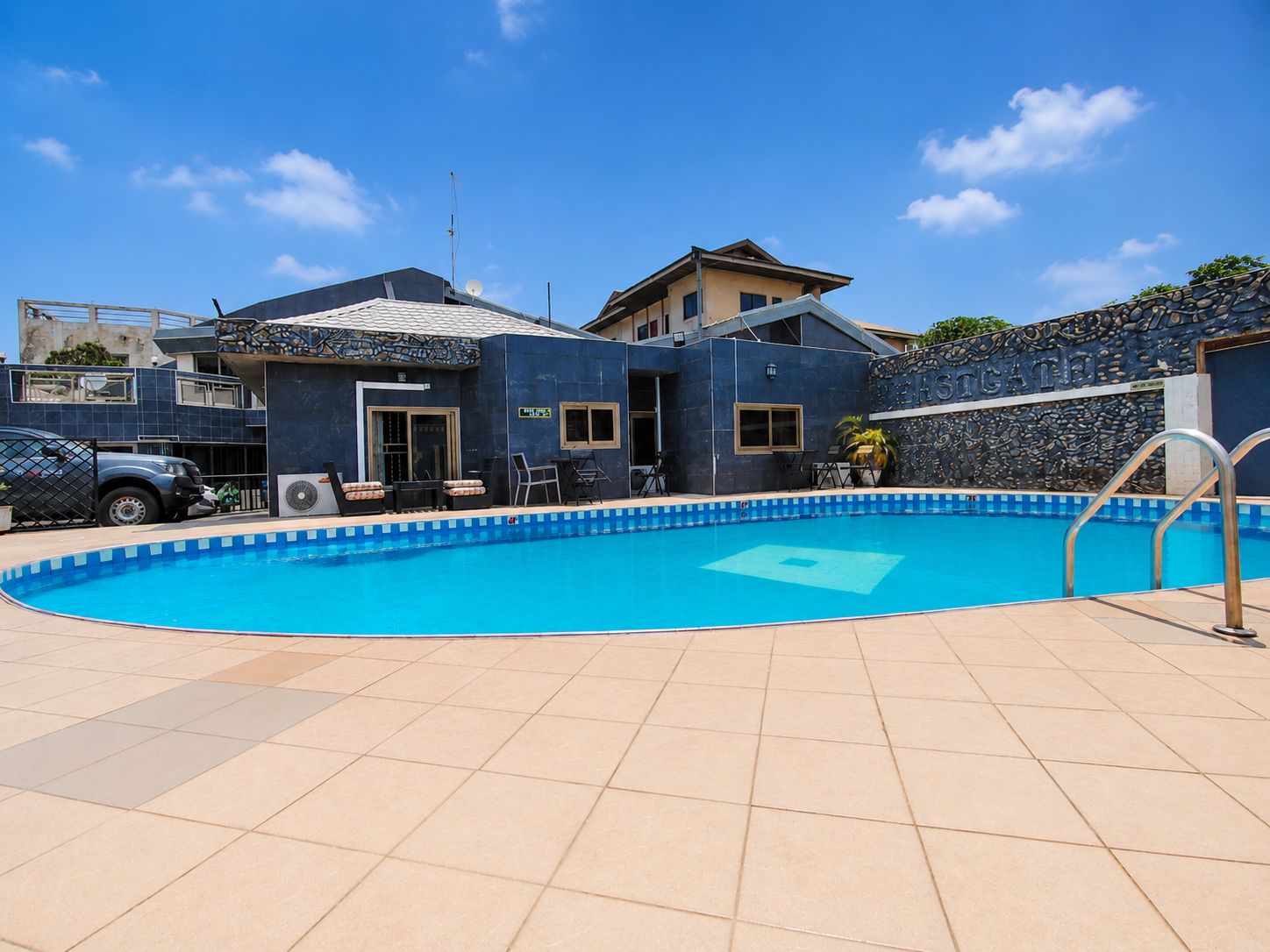 A swimming pool with a tiled deck, a covered area with chairs, and a stone wall with a sign.