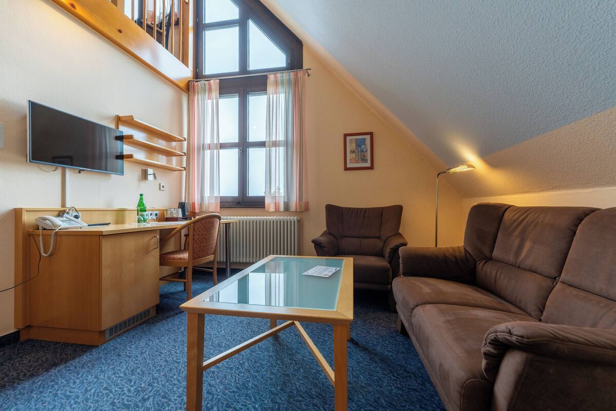 Una habitación de hotel con escritorio, sofá de cuero marrón, mesa de centro de vidrio, televisor montado en la pared y ventana con cortinas.