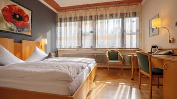 A hotel room with a bed, a desk, a chair, a radiator, and a window with curtains.