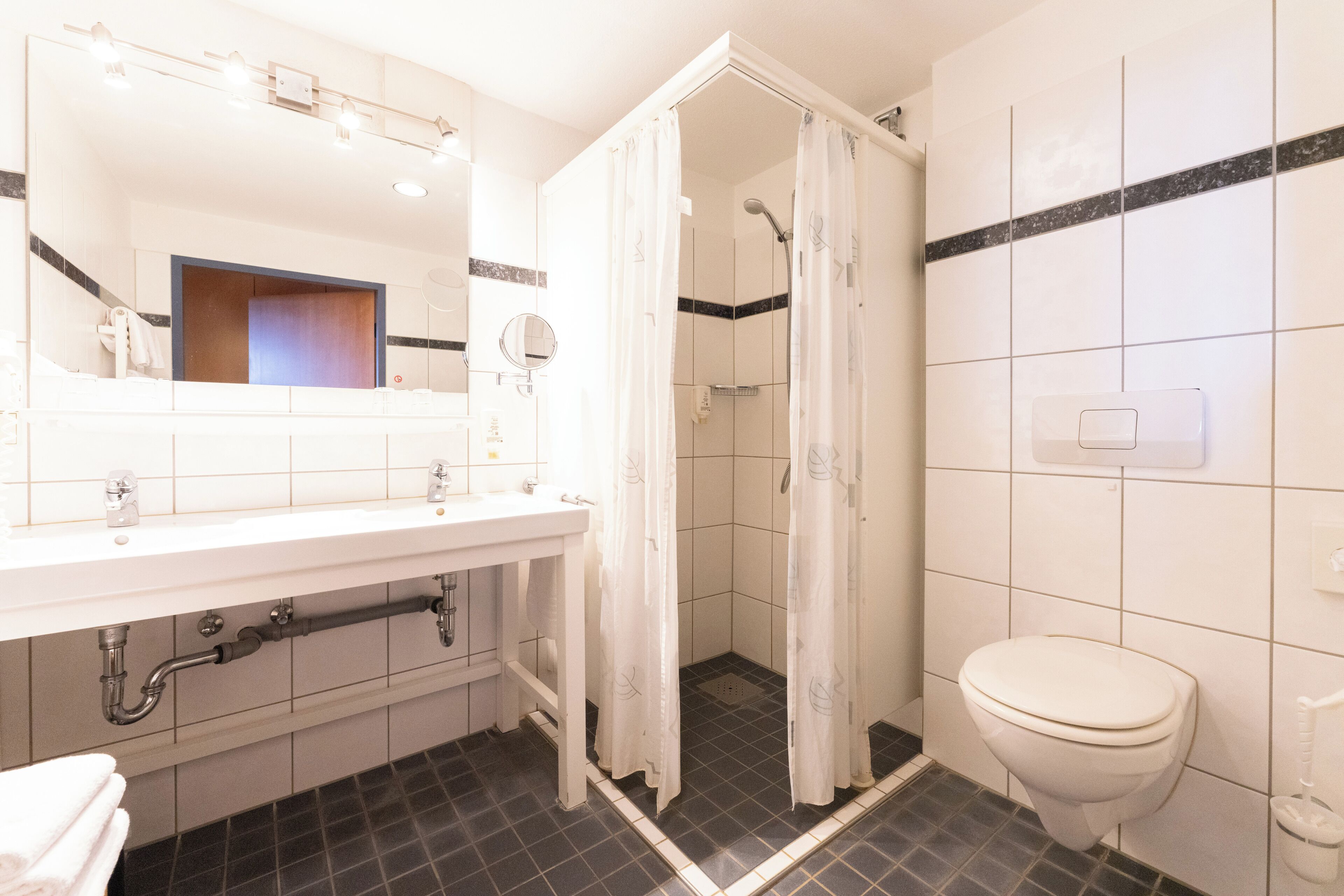 Un baño con lavamanos blanco, espejo e inodoro. Hay una cortina de ducha y piso de baldosas.