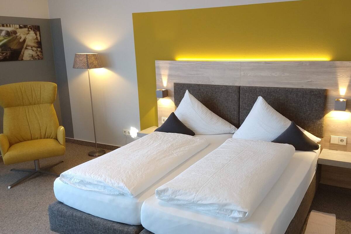 Una habitación de hotel con cama, dos almohadas, mesita de noche, lámpara y una silla.