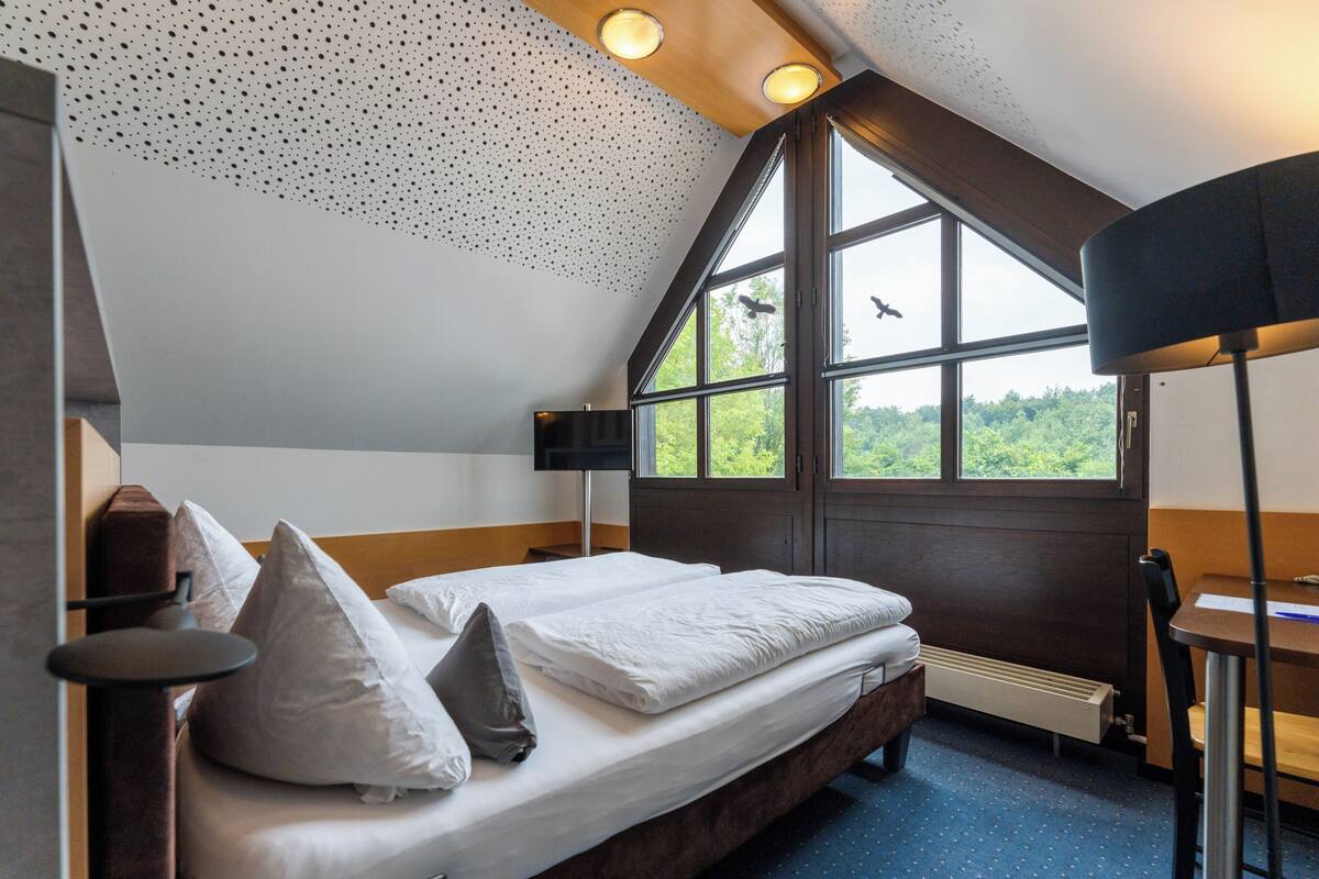 Un dormitorio con un ventanal grande, una cama con almohadas, un escritorio y una silla.