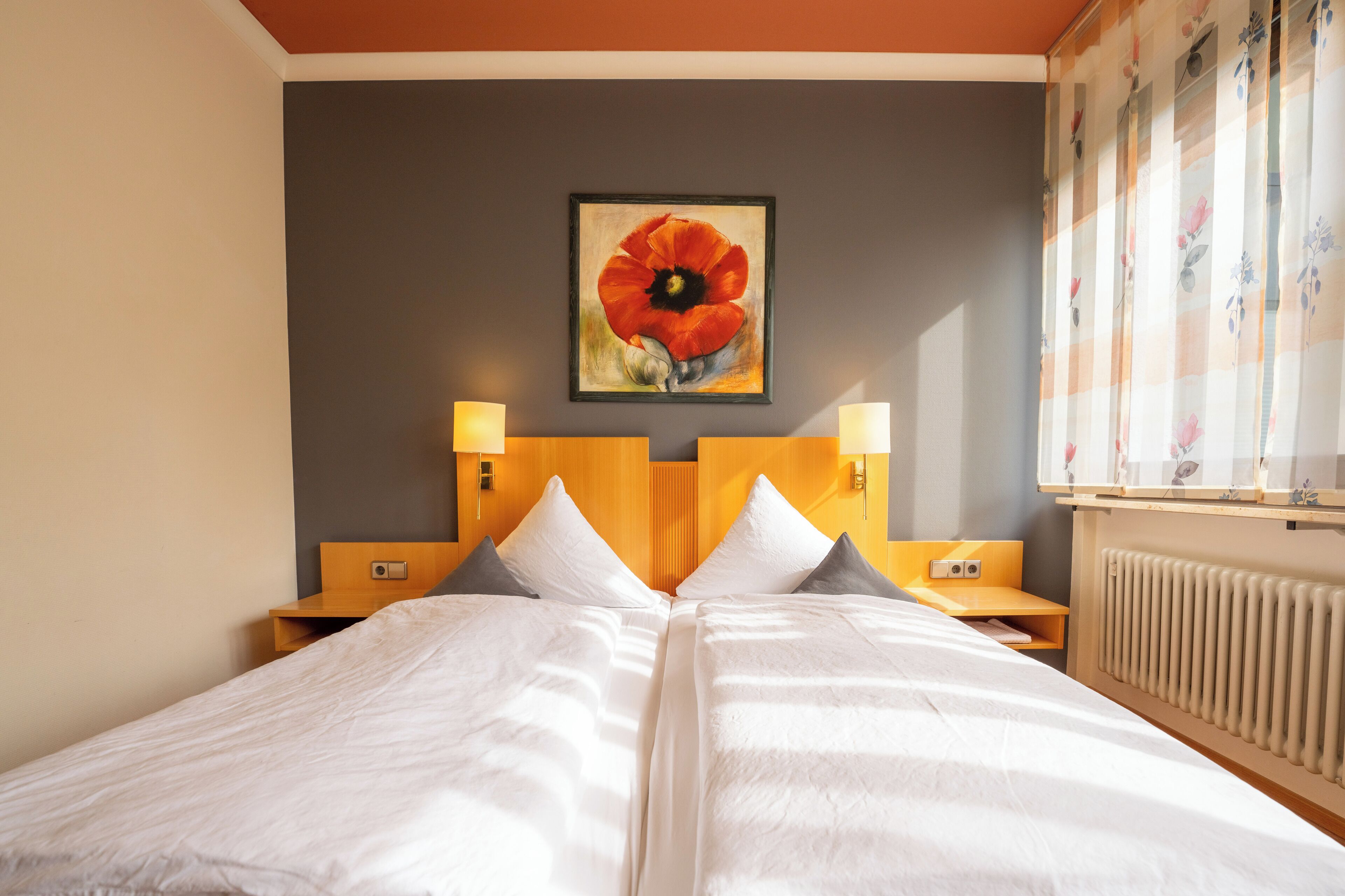 A double bed with white linens, two bedside tables with lamps, a poppy painting on the wall, and a window with floral curtains.
