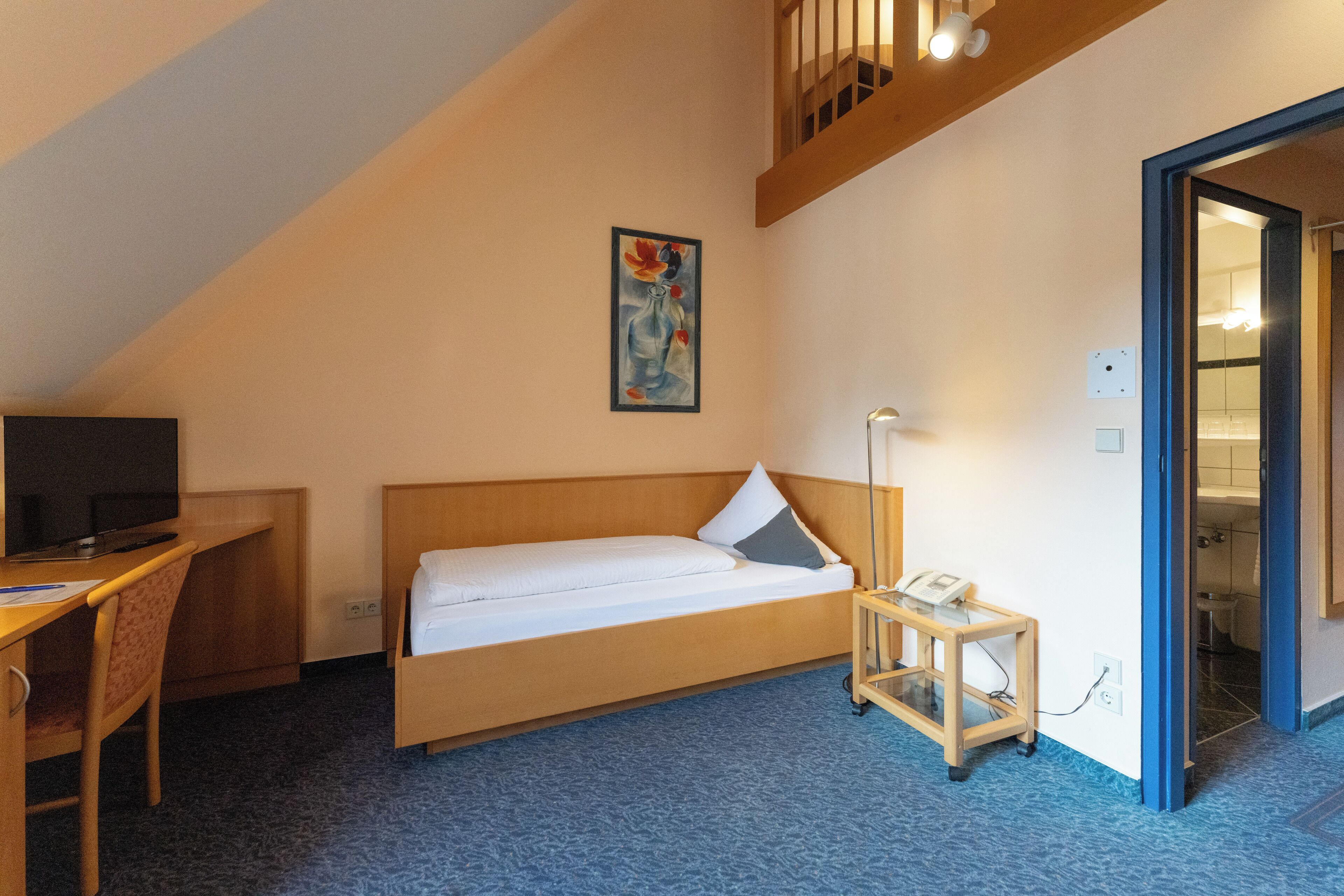 A small, single-bed room with a desk, a chair, a bedside table with a phone, and a painting on the wall.