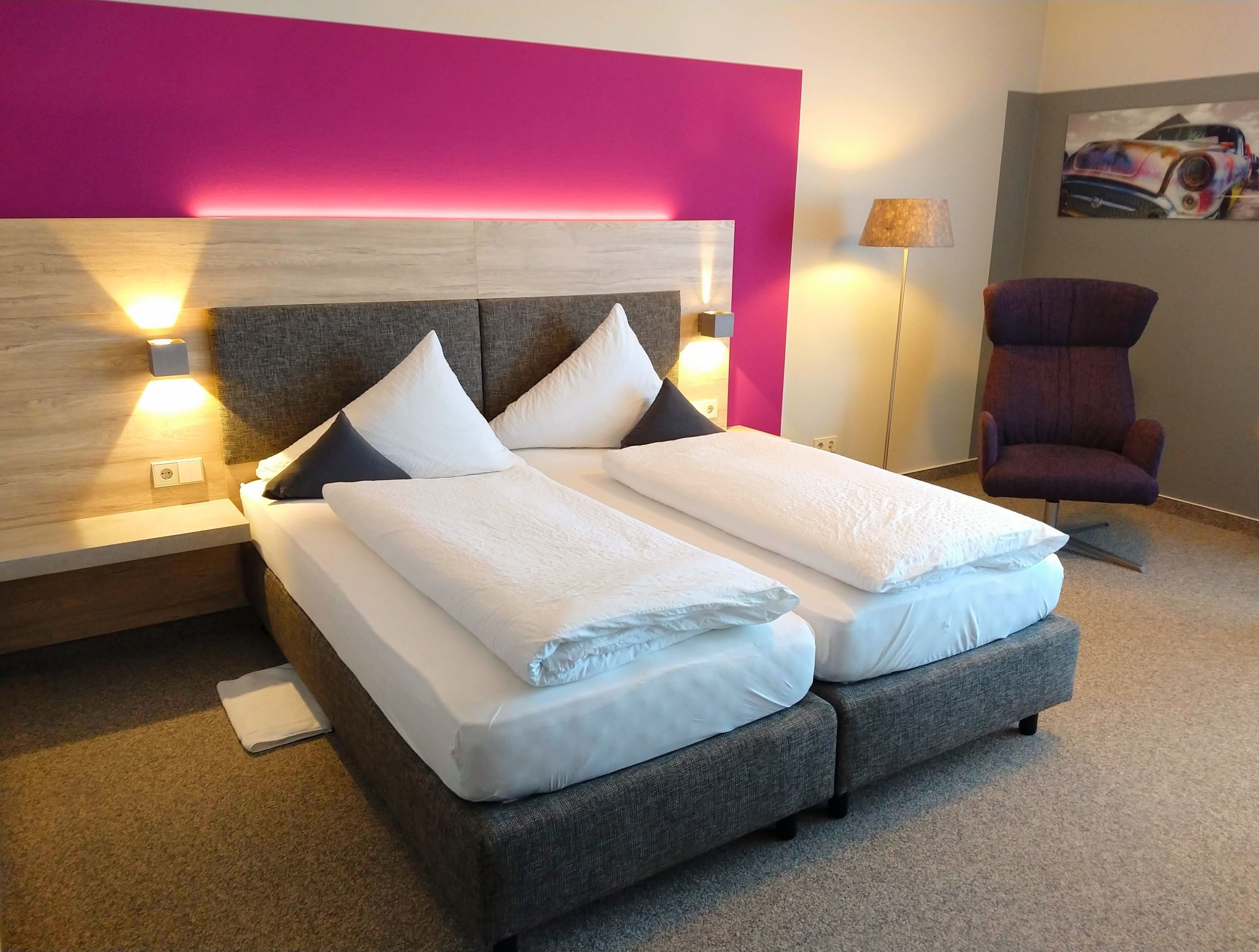 A modern hotel room with a large bed, a headboard with integrated lighting, a side table, a chair, and a painting on the wall.