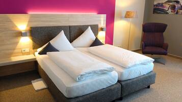 A modern hotel room with a large bed, a headboard with integrated lighting, a side table, a chair, and a painting on the wall.