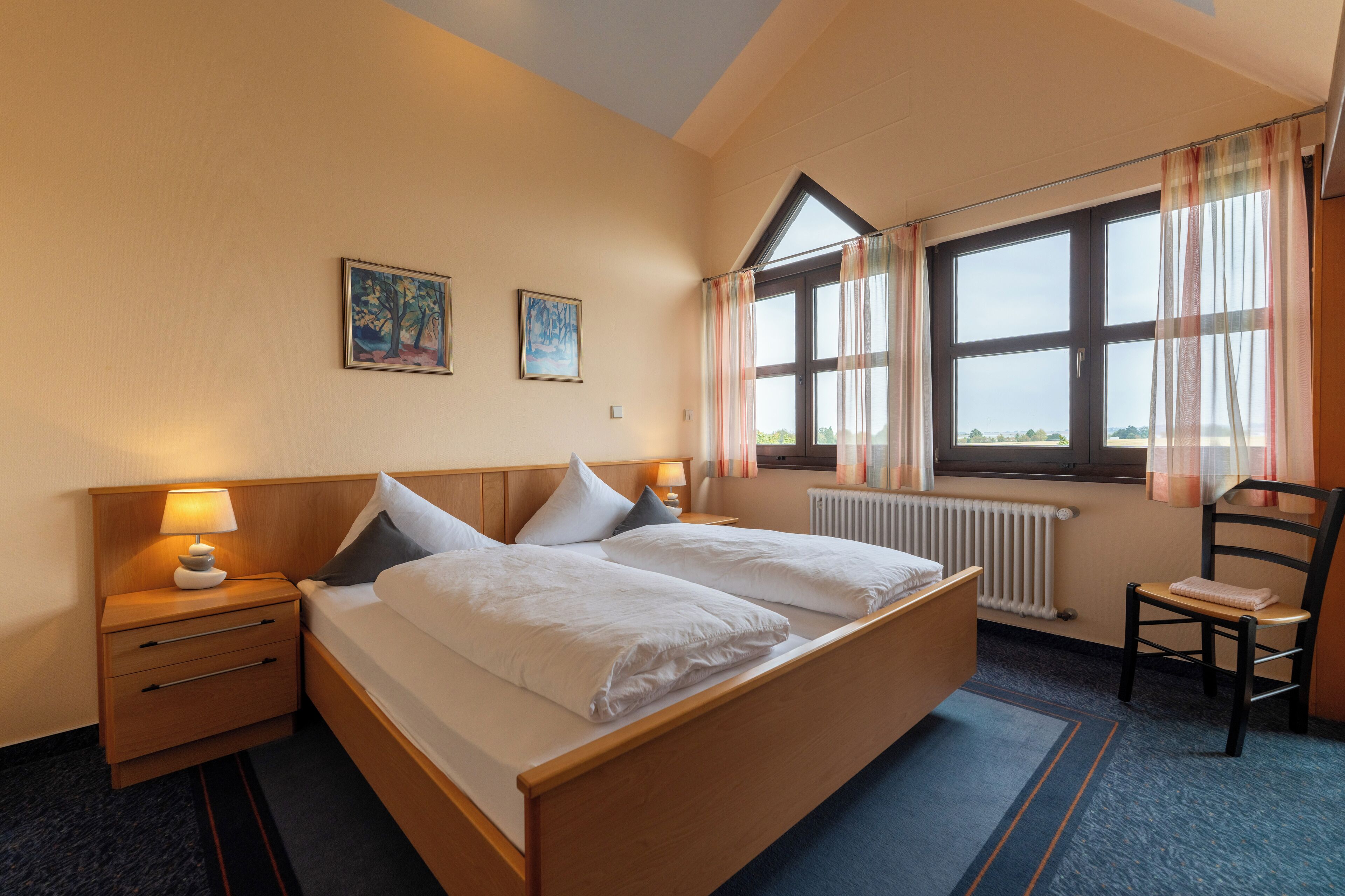 A hotel room with a large bed, two bedside tables with lamps, a chair, and two framed pictures on the wall.