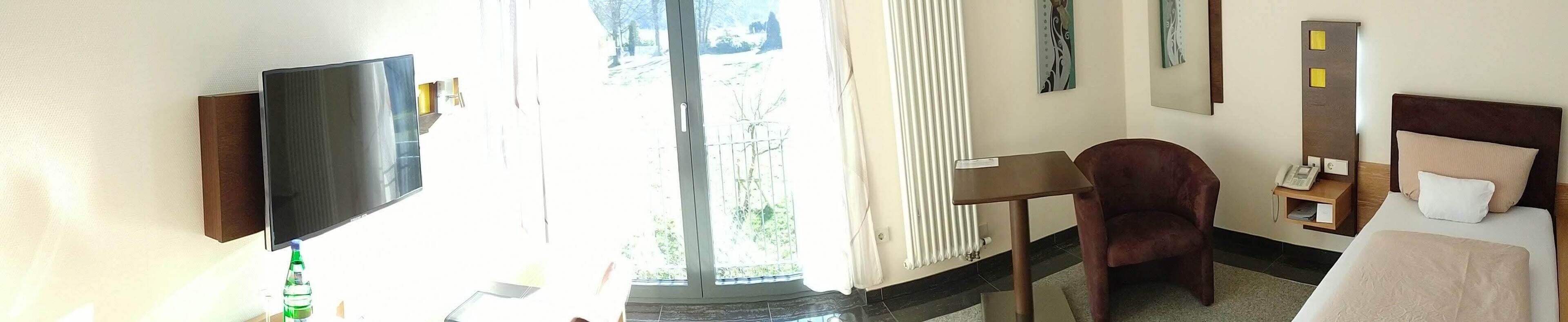 A hotel room with a flat-screen TV, a desk, a chair, a bed, and a window with a view of greenery.