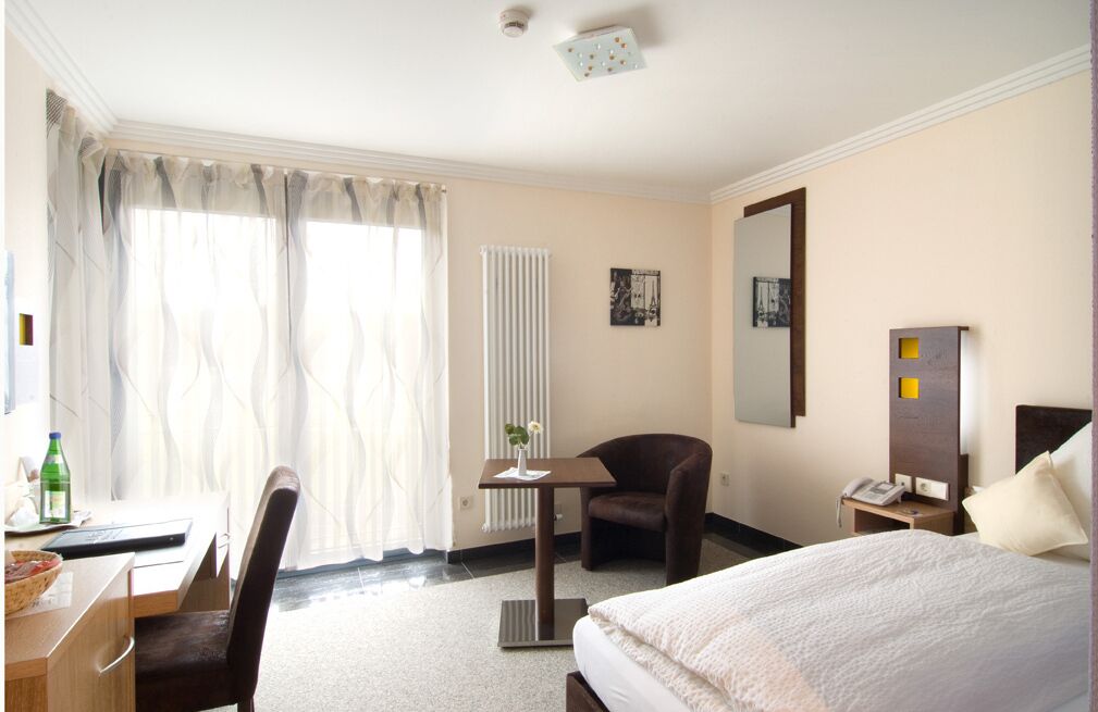 A hotel room with a bed, a desk with a chair, a small table with a vase, a chair, a bedside table with a lamp, a mirror, and a radiator.