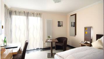 A hotel room with a bed, a desk with a chair, a small table with a vase, a chair, a bedside table with a lamp, a mirror, and a radiator.