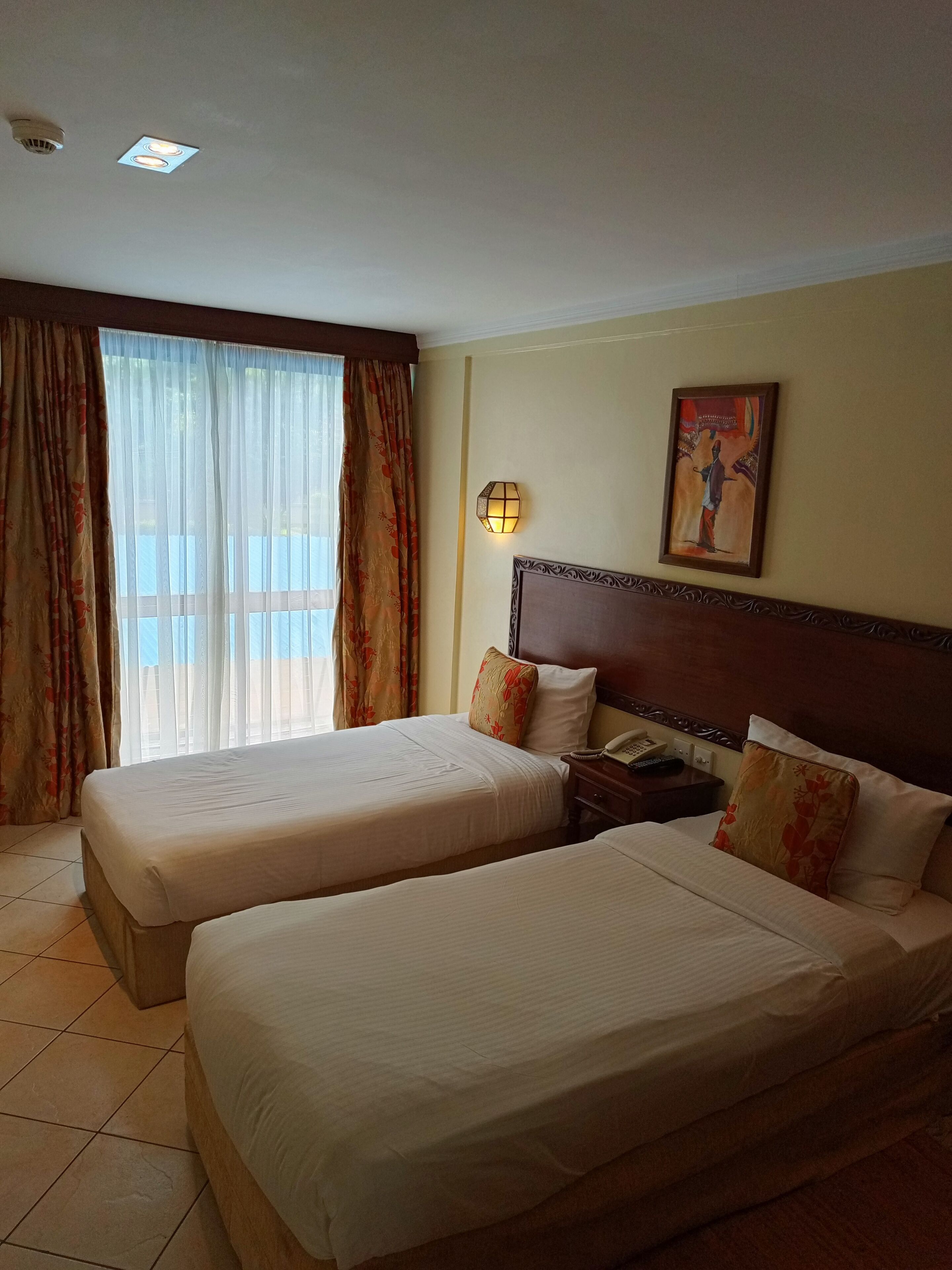 A hotel room with two beds, a nightstand, a wall-mounted lamp, and a painting.