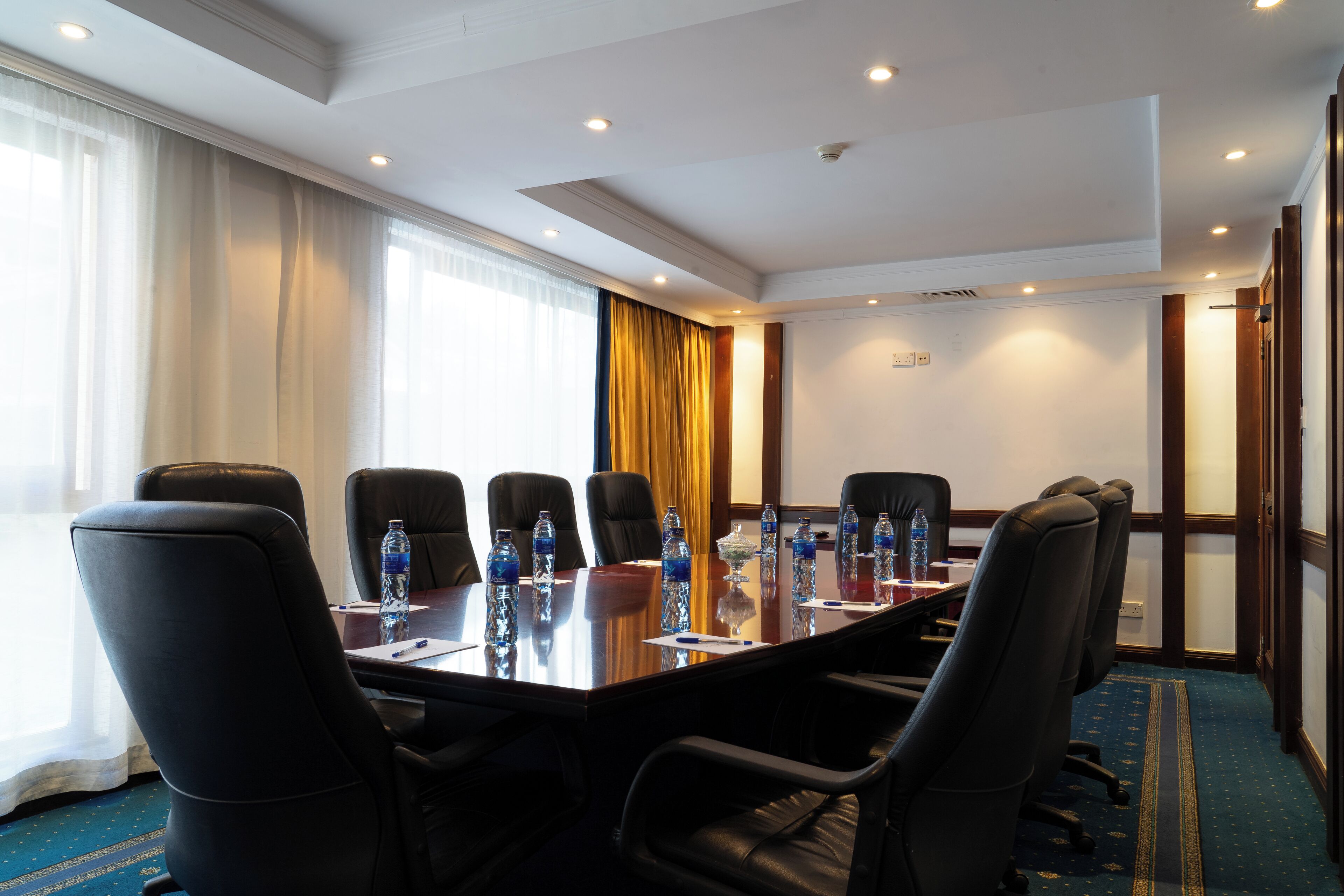 A conference room with a long table, black leather chairs, water bottles, and a blue carpet.