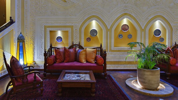 Un interior tradicional con decoraciones ornamentales en las paredes, un sofá rojo con numerosos cojines, una silla de madera y una mesa de centro.