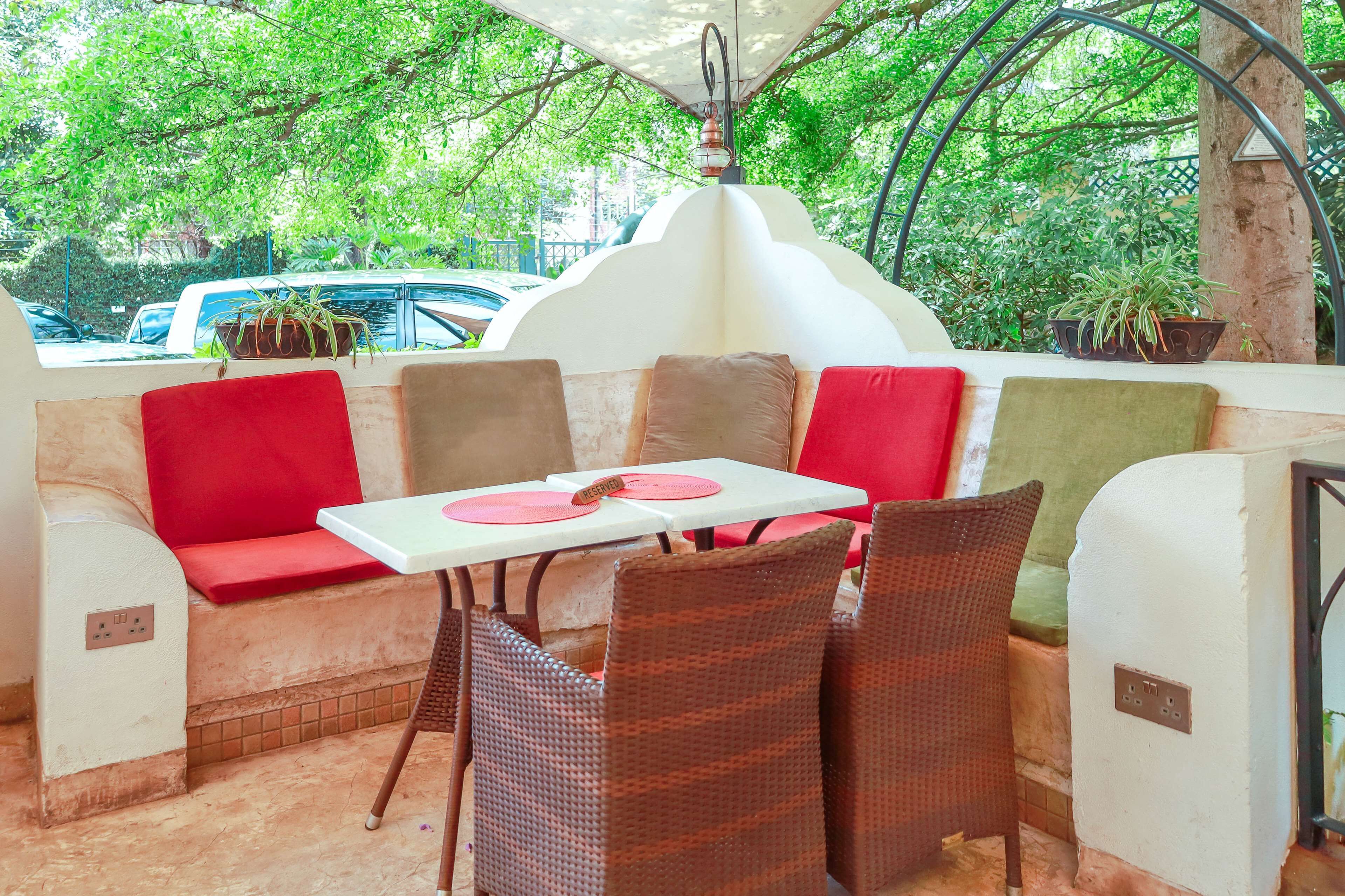 Un área de estar al aire libre con sillas de mimbre y cojines rojos, una mesa blanca y plantas en macetas.