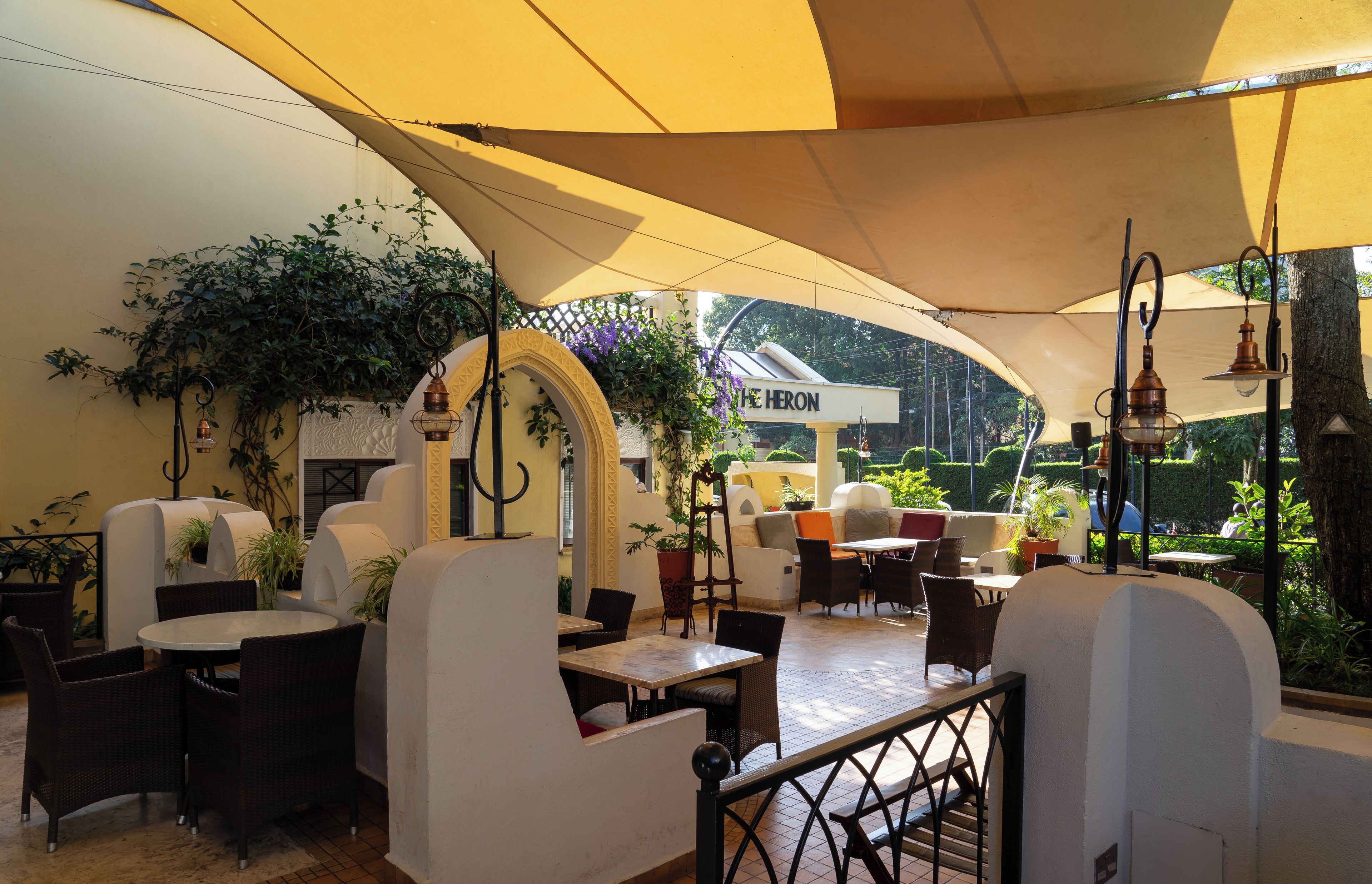 An outdoor café with round tables and chairs, potted plants, and a canopy providing shade.