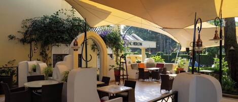 An outdoor café with round tables and chairs, potted plants, and a canopy providing shade.