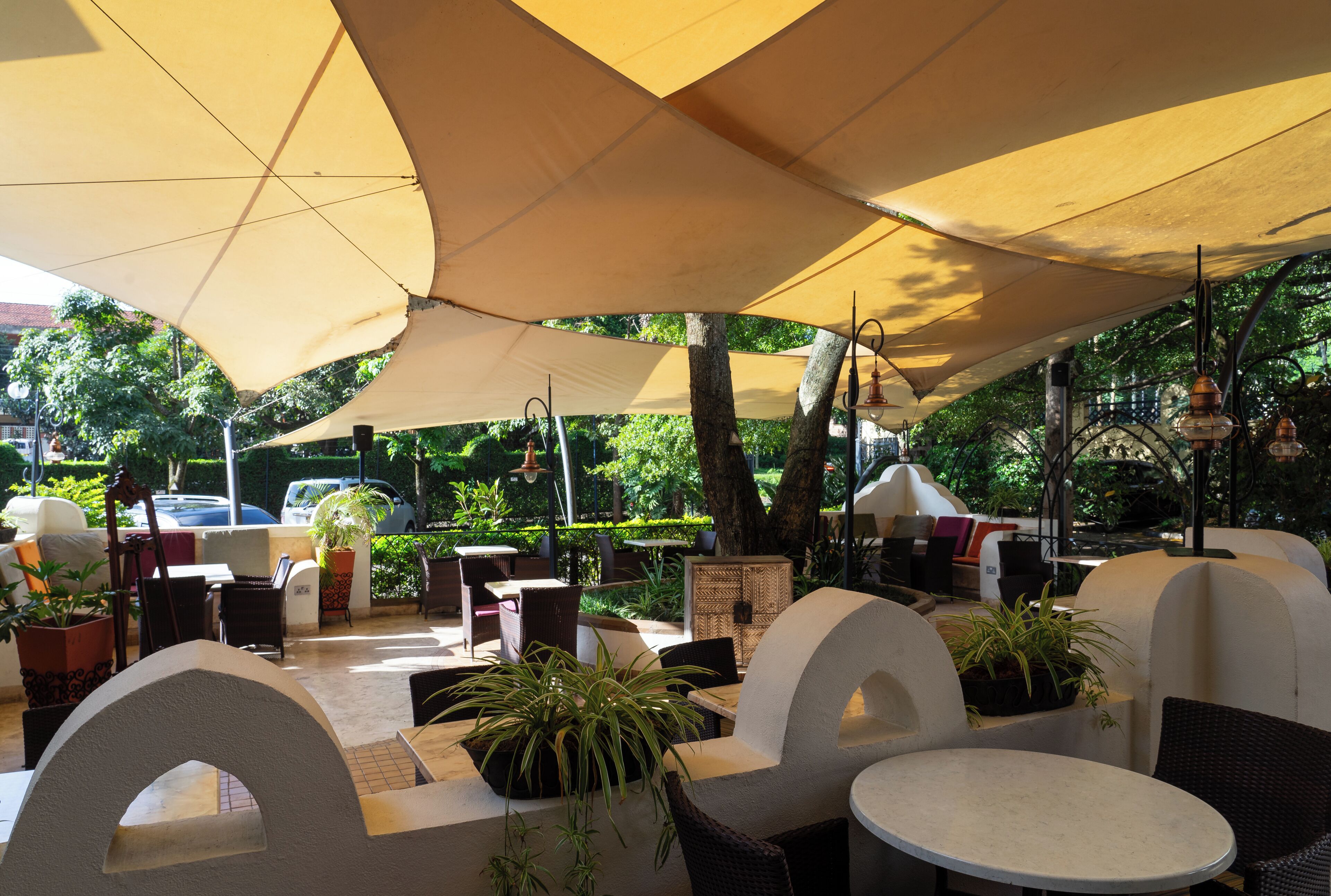 An outdoor seating area with round tables and chairs, shaded by large umbrellas, surrounded by potted plants and trees.
