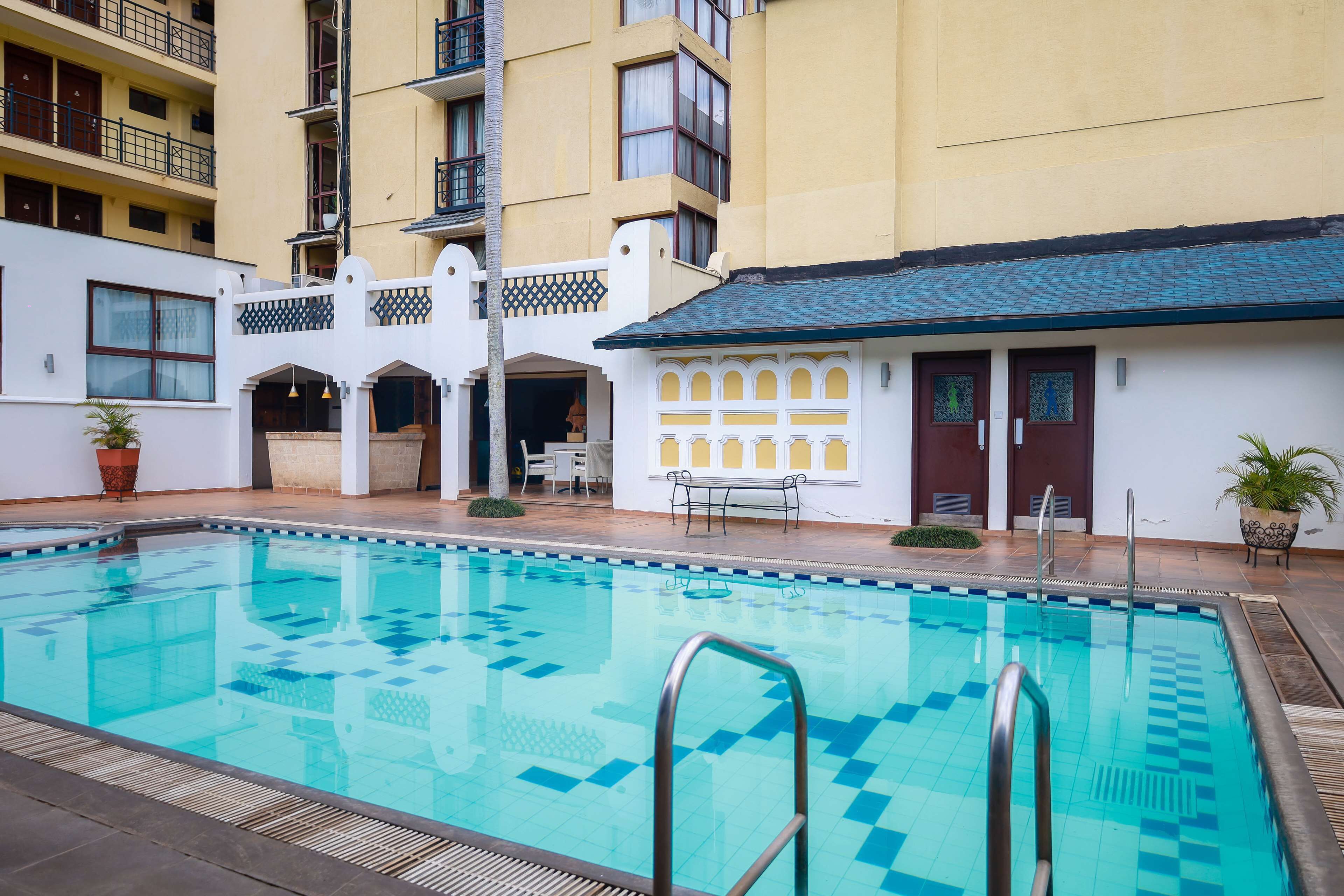 Une piscine avec une terrasse carrelée, un bâtiment blanc doté de balcons et un petit espace extérieur aménagé pour s’asseoir.
